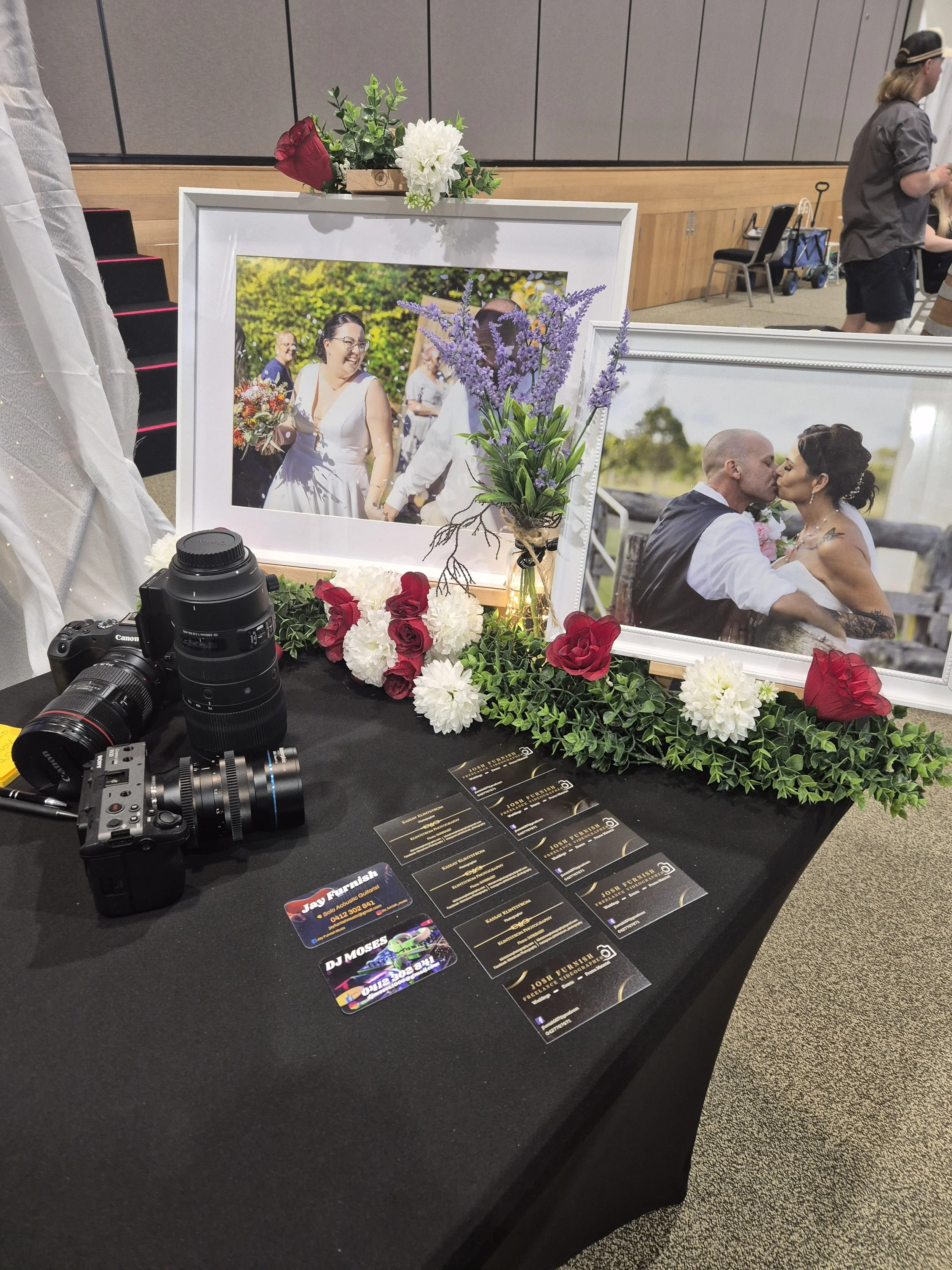 Display table with two framed wedding photos, floral arrangements, business cards, and camera equipment at an event.