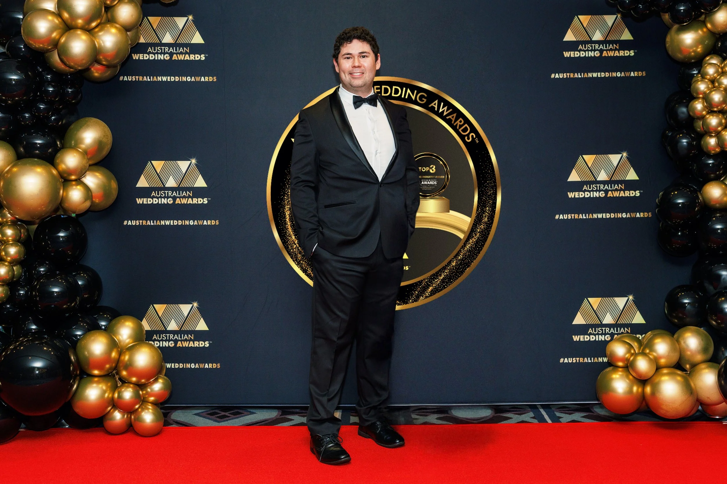 A man in a tuxedo standing on a red carpet at the Australian Wedding Awards, with a backdrop decorated with gold and black balloons and the event logo.