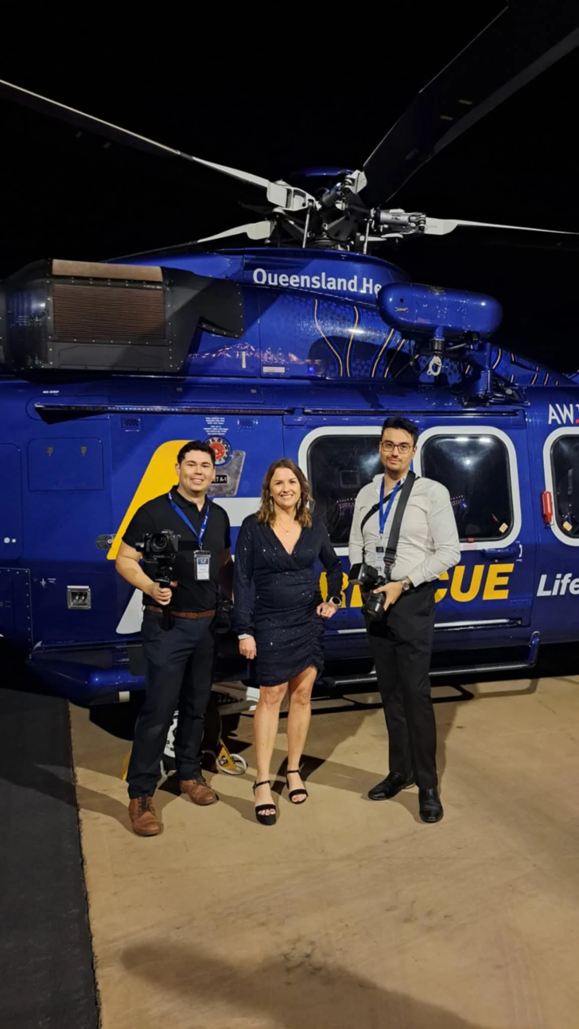 Three people standing in front of a blue rescue helicopter at night. The helicopter has 'Queensland H' and 'RESCUE' written on it. The group includes two men, one woman, all dressed in formal or semi-formal attire, with the men holding cameras.