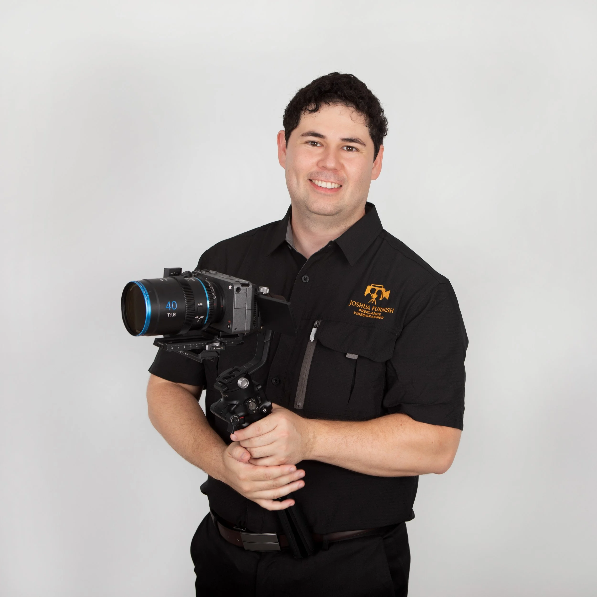A man in a black shirt holding a camera on a stabilizer, standing against a plain light gray background.