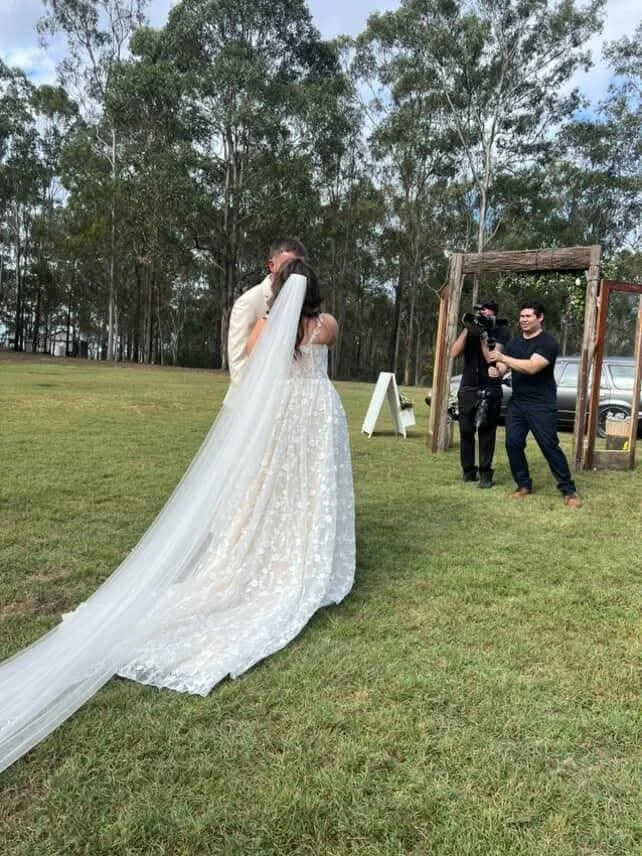 A bride in a wedding dress and veil kissing a groom in an outdoor setting with photographers capturing the moment.