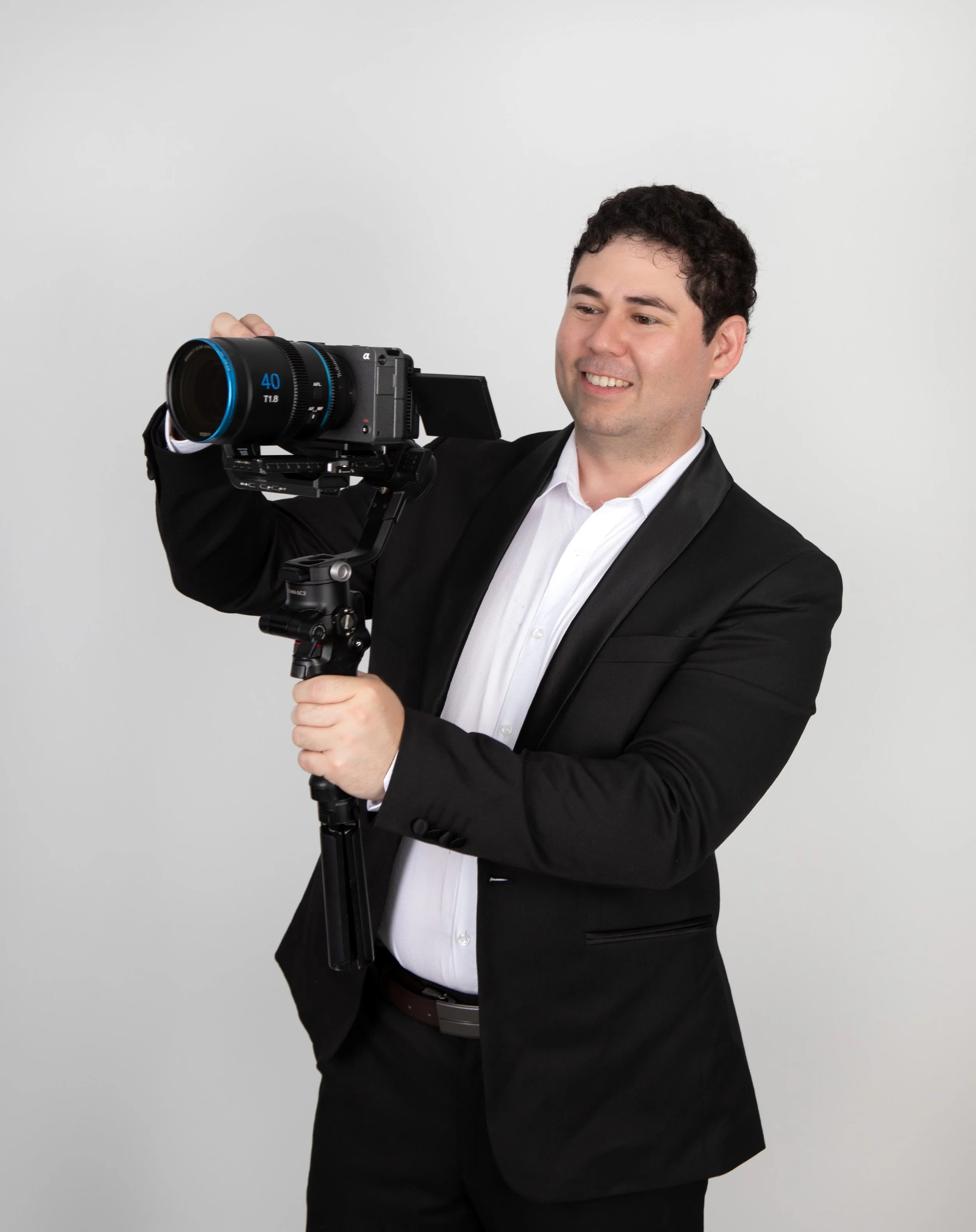 A man in a black suit and white shirt holding a professional camera on a tripod, smiling.