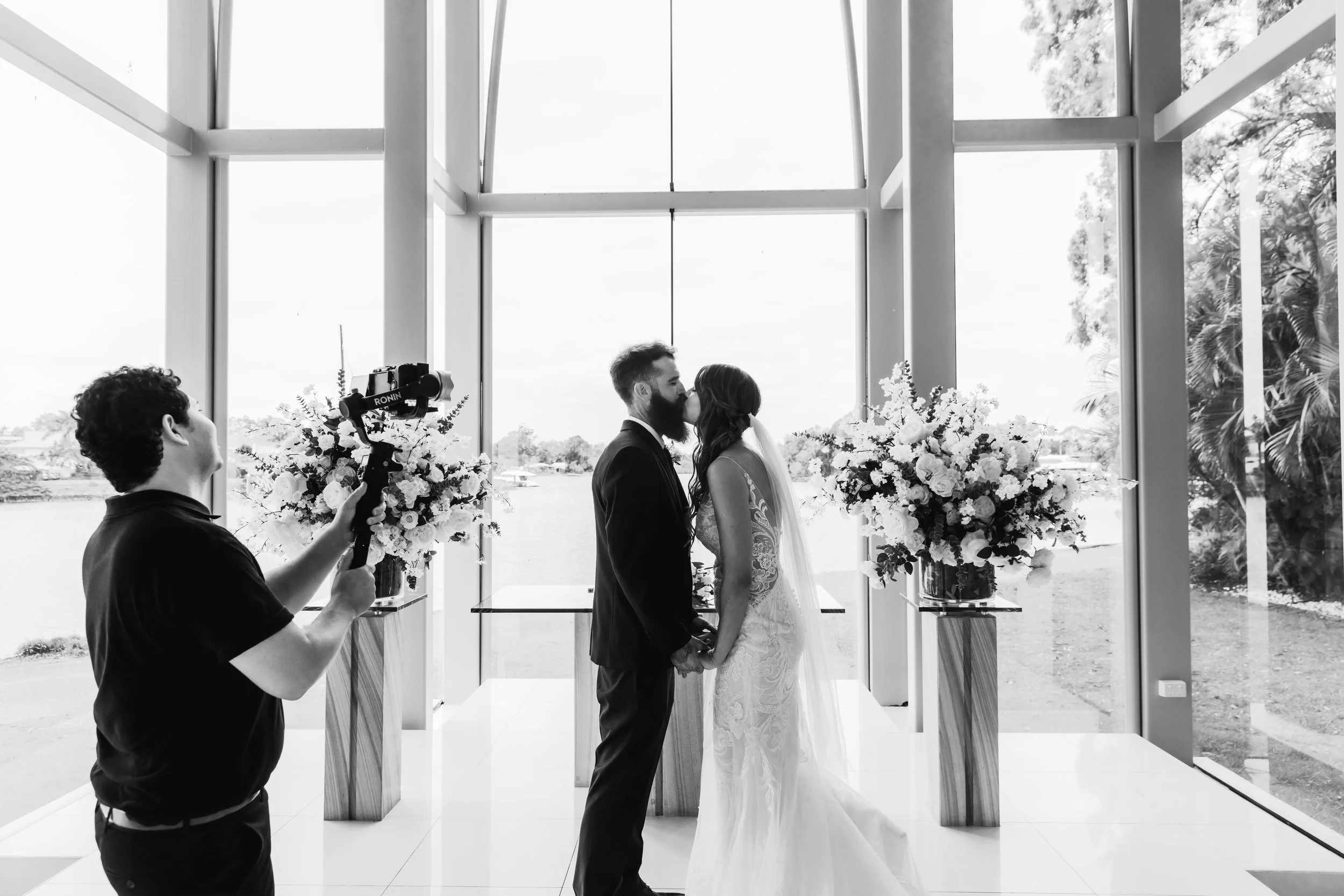 A black and white photo of a bride and groom kissing during their wedding ceremony inside a modern glass-walled building with large windows and floral arrangements on either side.