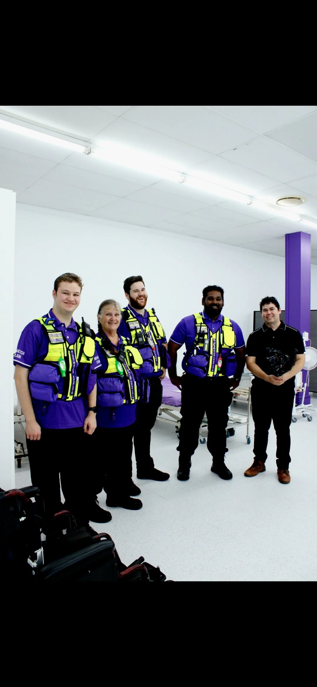 Five paramedics and a man holding equipment standing in a hospital room with white walls, bright lighting, and medical beds.