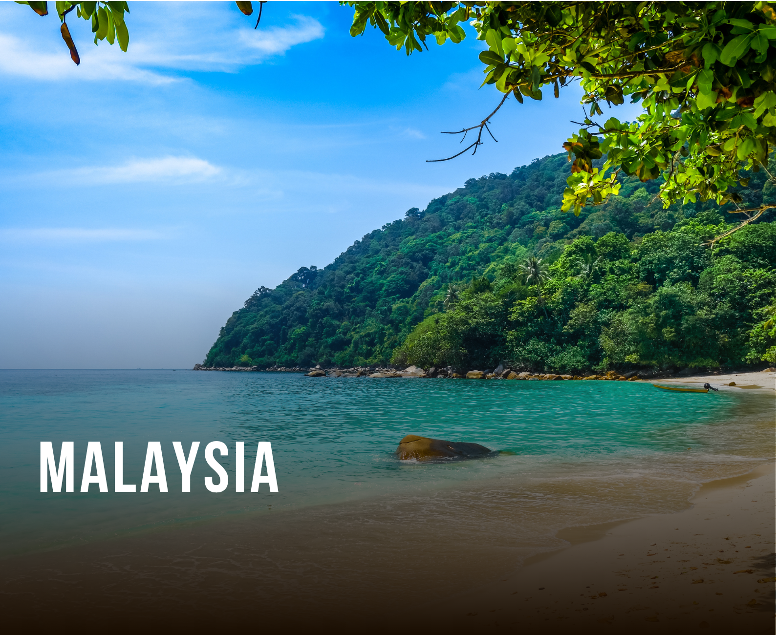 Tropical beach in Malaysia with clear turquoise water, sandy shore, lush green hillside, and trees overhanging the beach, under a blue sky with some clouds.