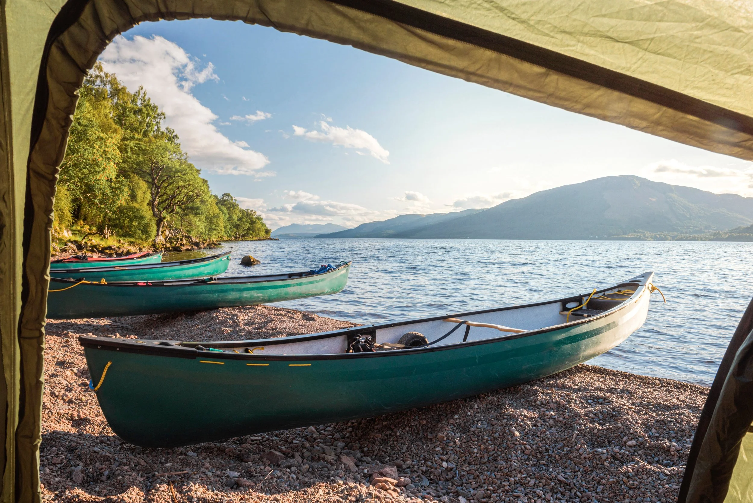 View from inside a tent overlooking a lakeshore with boats and mountains, with a bright blue sky and scattered clouds.