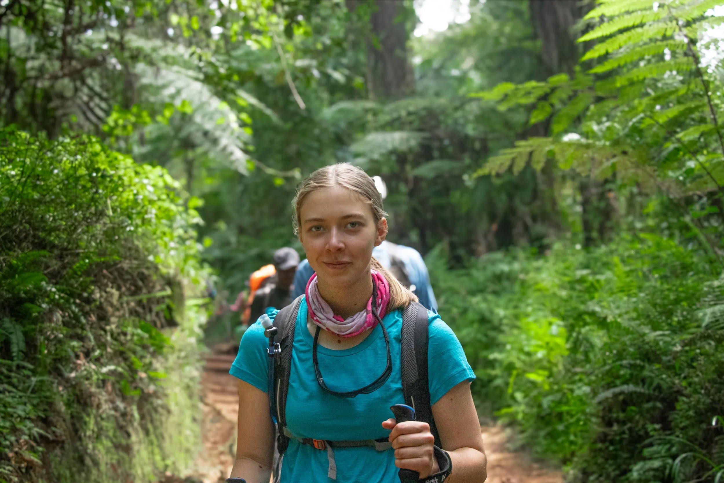 A young woman with braided hair and a blue shirt is hiking on a forest trail, carrying a backpack and holding hiking poles. She is smiling and looking ahead as she walks through lush green trees and foliage.