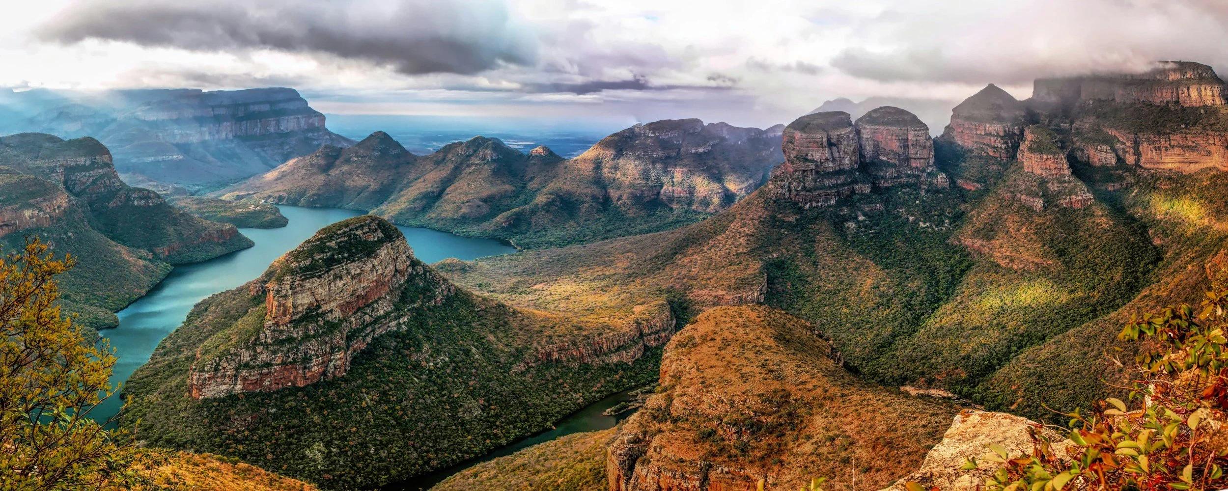 A panoramic view of a canyon with steep cliffs, a winding river, and rolling hills under a cloudy sky.