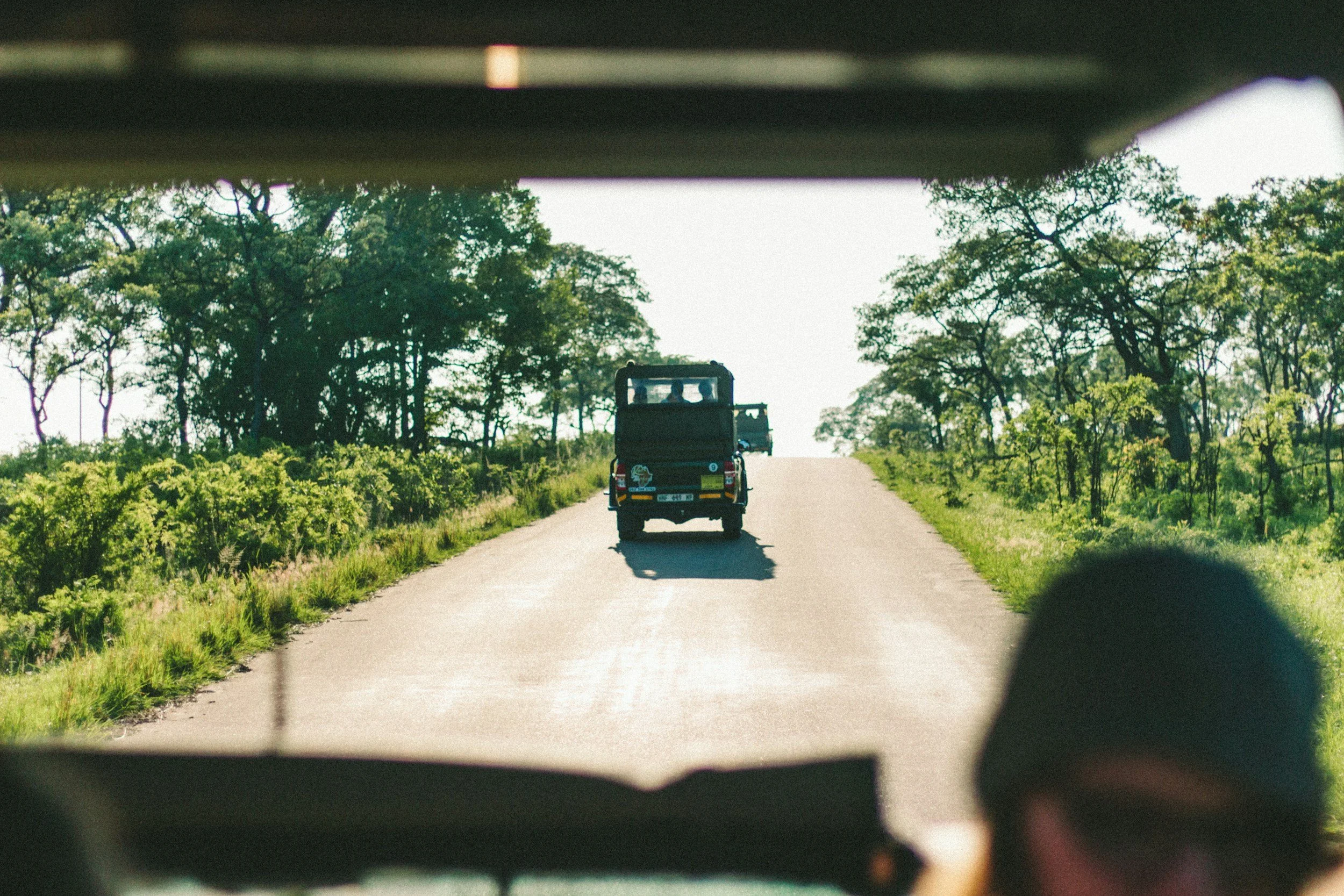 A view through the rear window of a vehicle showing a black off-road vehicle driving on a rural road surrounded by green trees and bushes, under a bright sky.