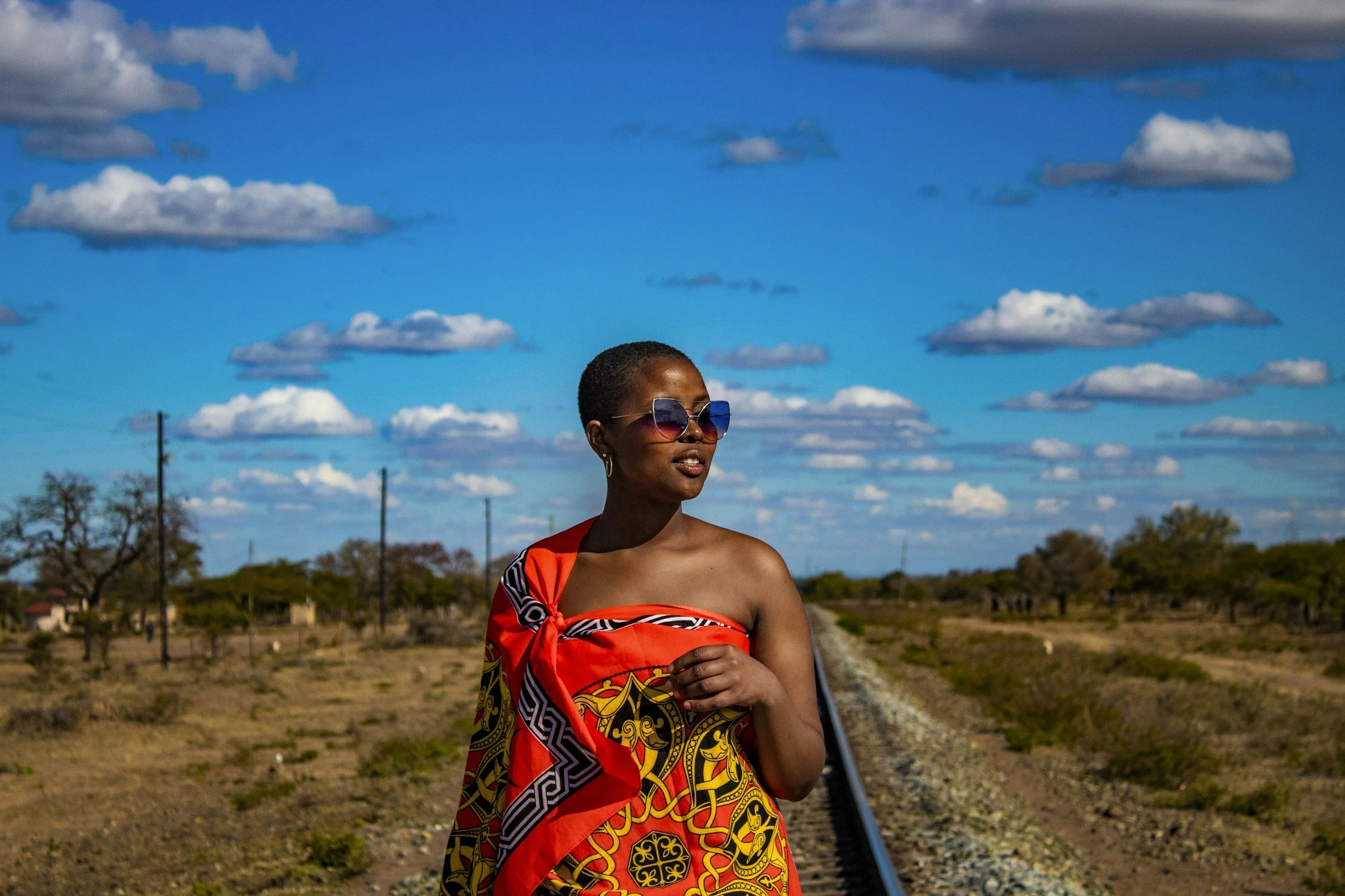 A woman with short hair wearing sunglasses and a vibrant, patterned dress stands outdoors near train tracks under a bright blue sky with scattered clouds.