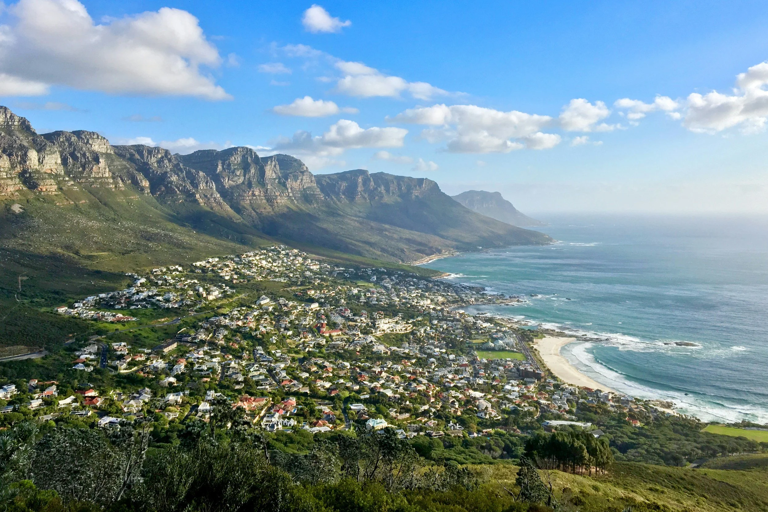 A coastal town with houses stretching along the shoreline, set against steep rocky mountains under a partly cloudy sky.