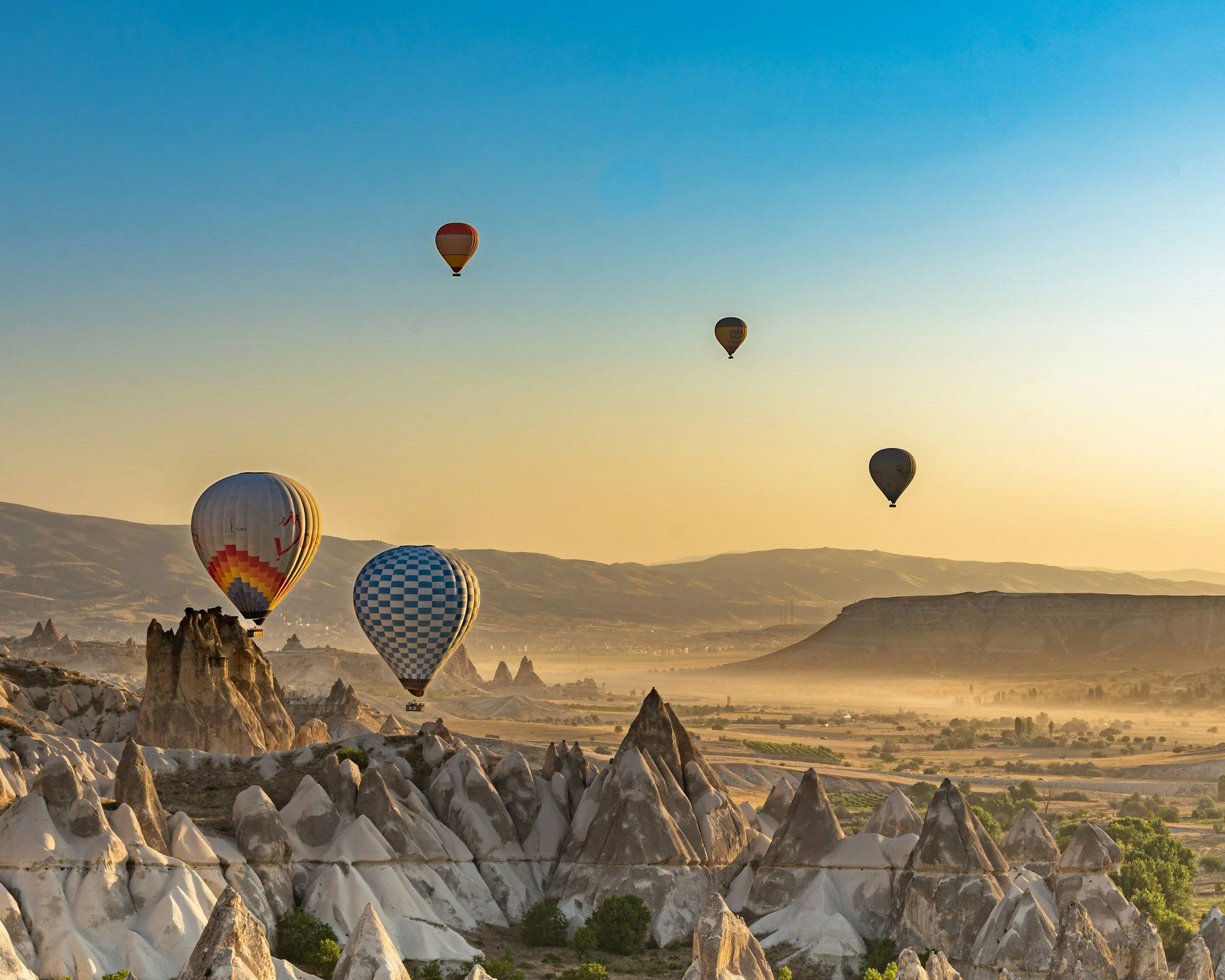 Multiple hot air balloons floating over unique rock formations and a scenic landscape during sunrise or sunset.
