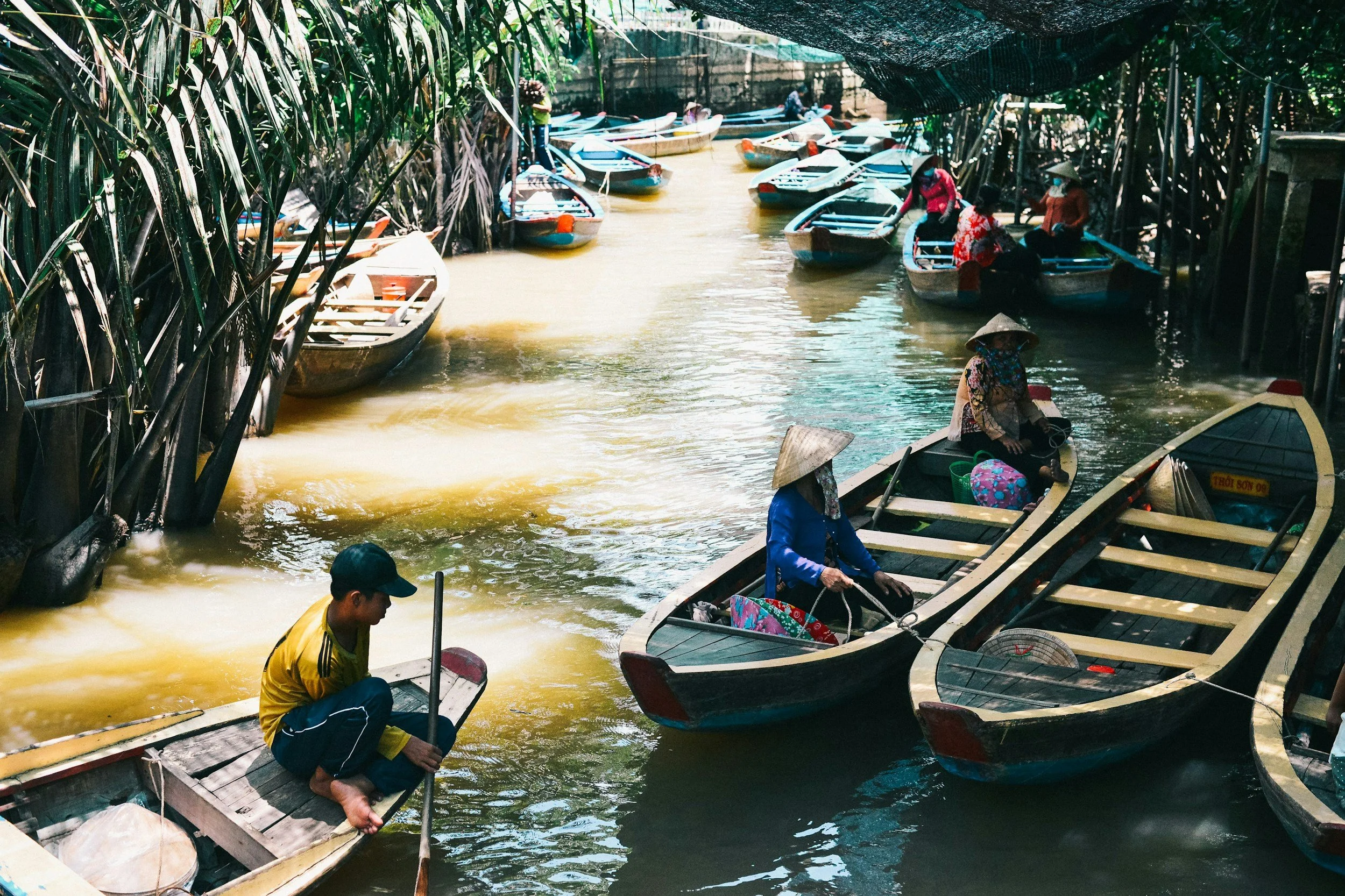 People in traditional hats and clothing riding small boats along a narrow, muddy river lined with tall green plants and trees, some people preparing or adjusting boats, under a shaded canopy.