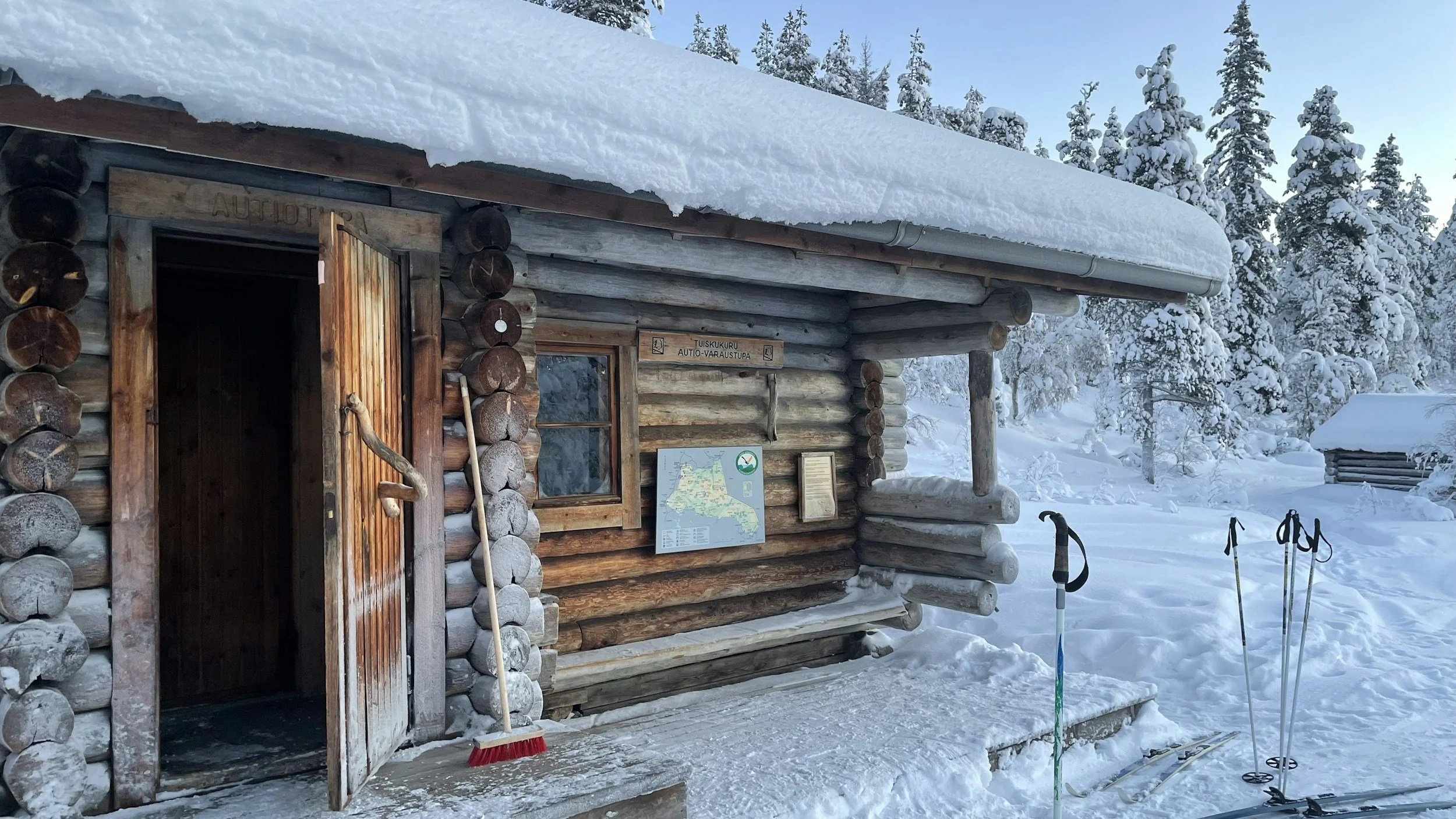 A rustic log cabin in a snowy landscape surrounded by snow-covered trees, with ski equipment and a broom outside.