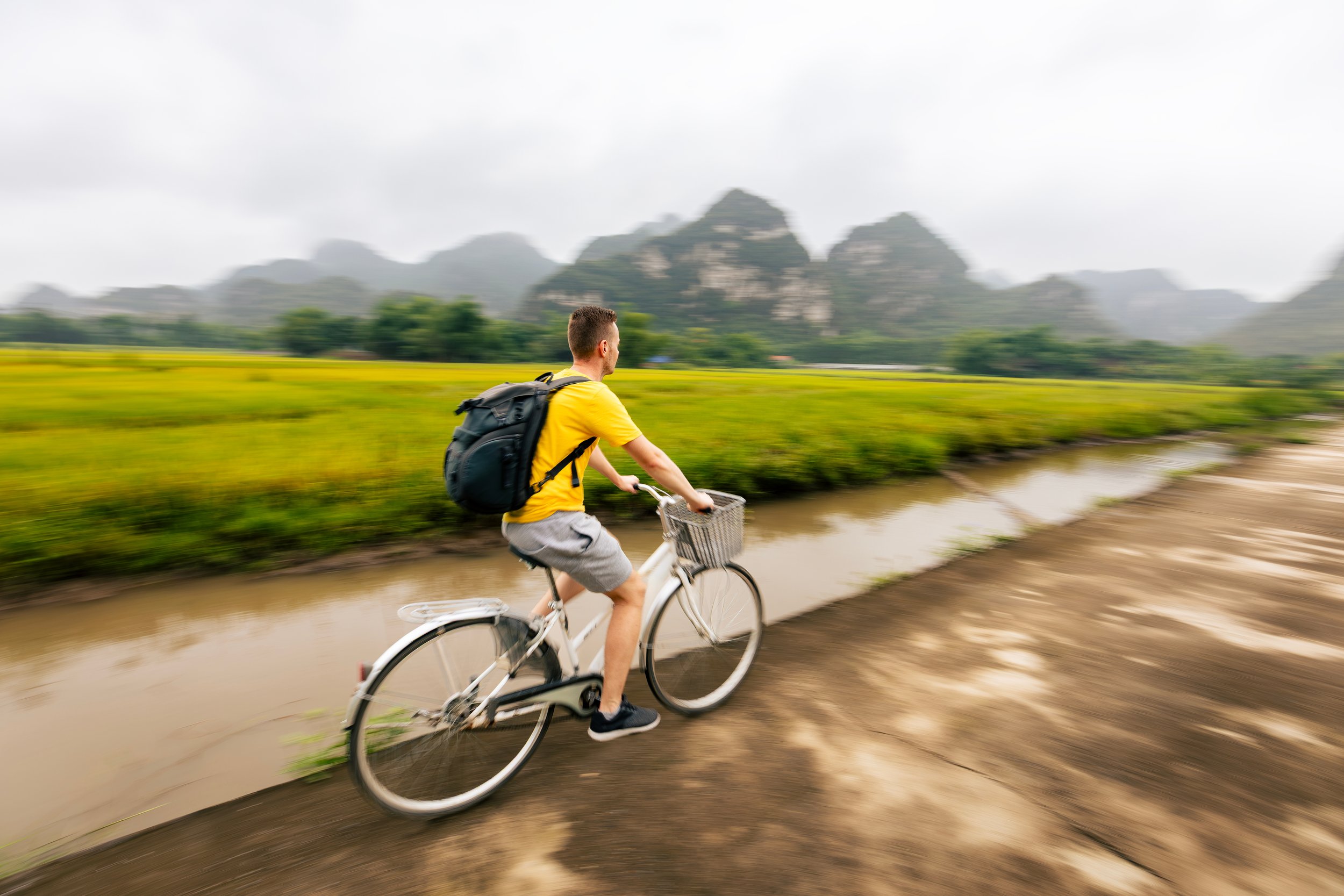 A man riding a bicycle on a dirt path through green fields with karst mountains in the background, wearing a yellow shirt and carrying a backpack.