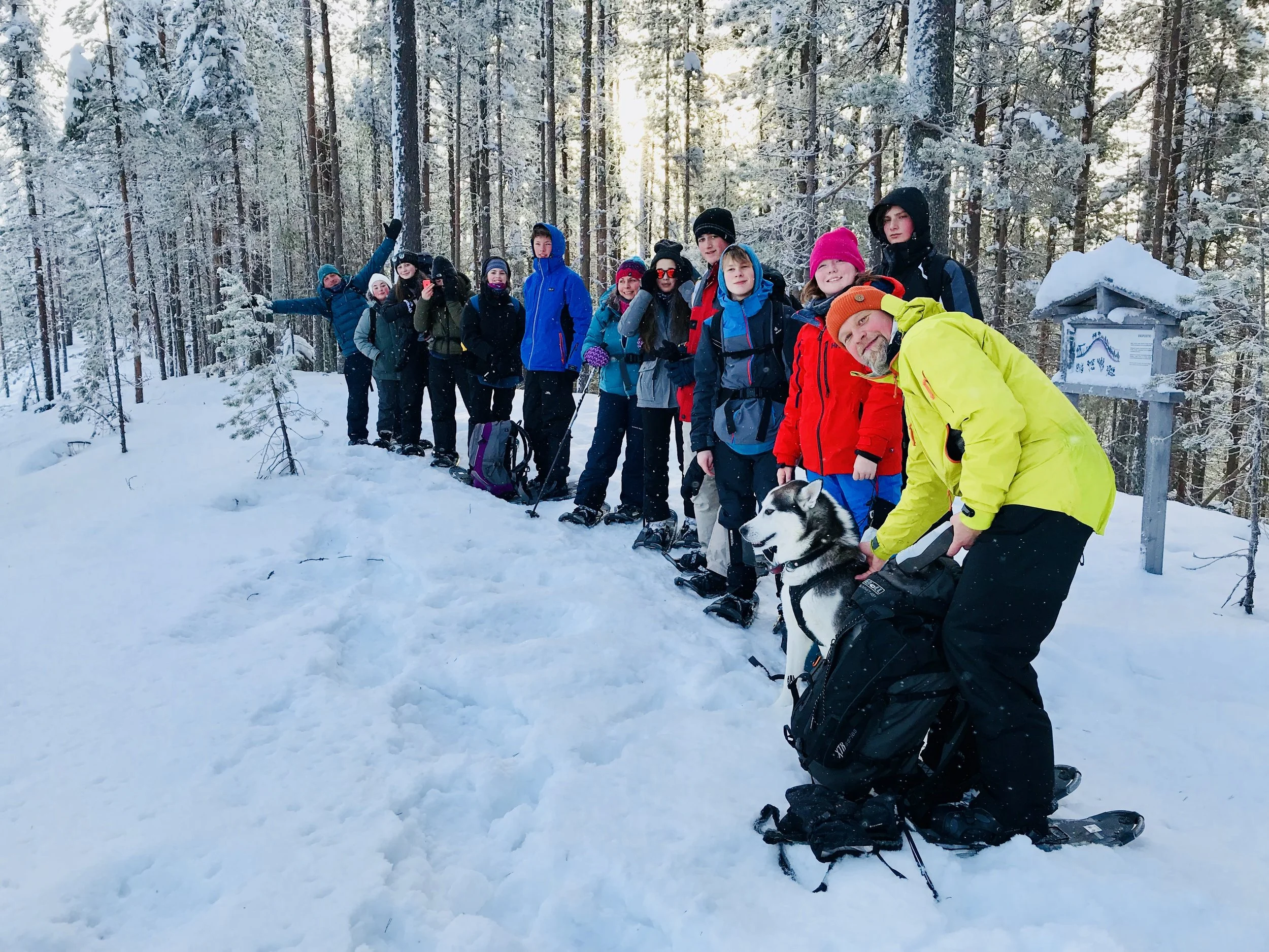 A group of people in winter clothing and snow gear standing in a snowy forest with trees covered in snow, smiling and posing for a photo. One person is kneeling in front with a dog.