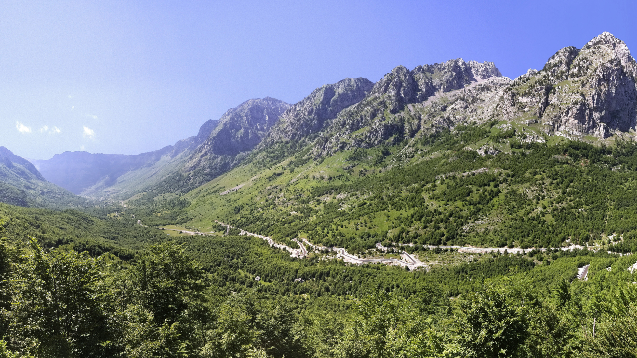 Green mountains with rocky peaks under a blue sky, winding roads through lush greenery in a valley.