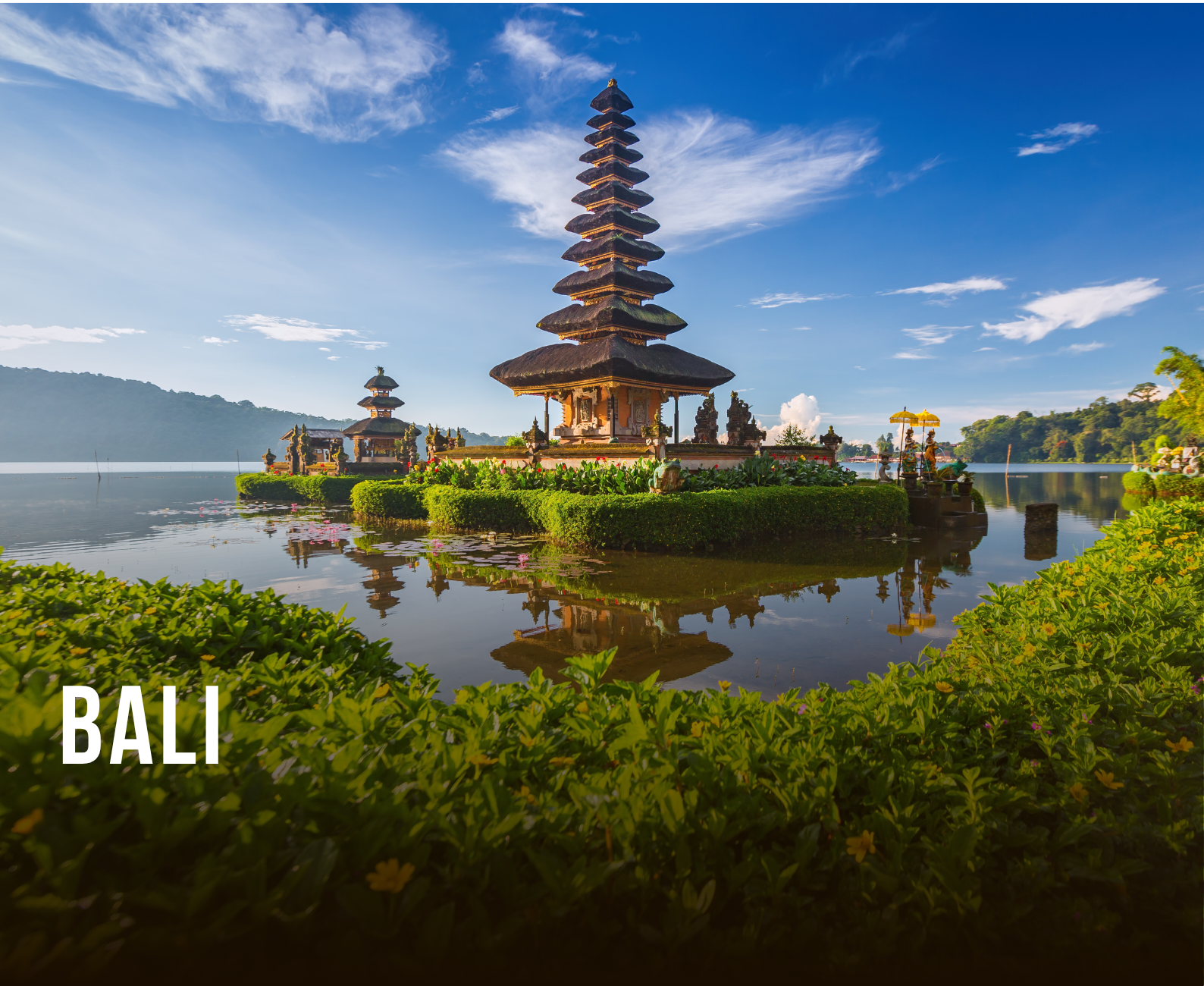 A traditional Balinese temple with multiple tiers, situated on a small island in a lake, surrounded by lush green vegetation and reflected in the water, under a bright blue sky.