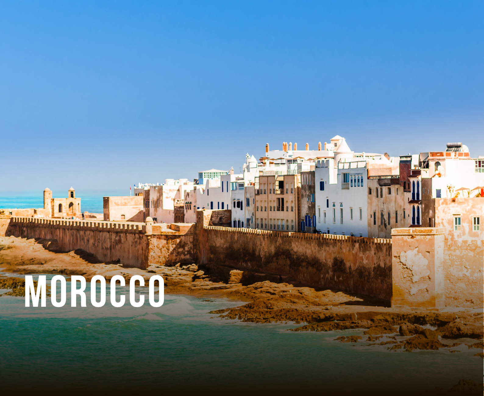 A coastal view of historic Moroccan buildings with a stone wall along the shoreline, under a clear blue sky in Morocco.