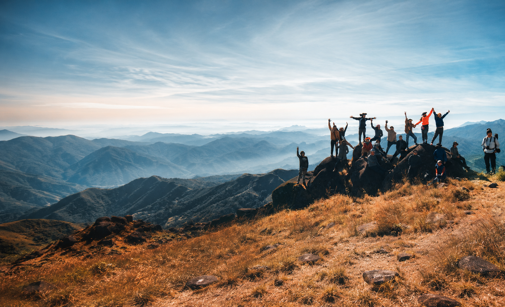 Group of people hiking and celebrating on a mountain ridge with panoramic view of mountain ranges and sky.
