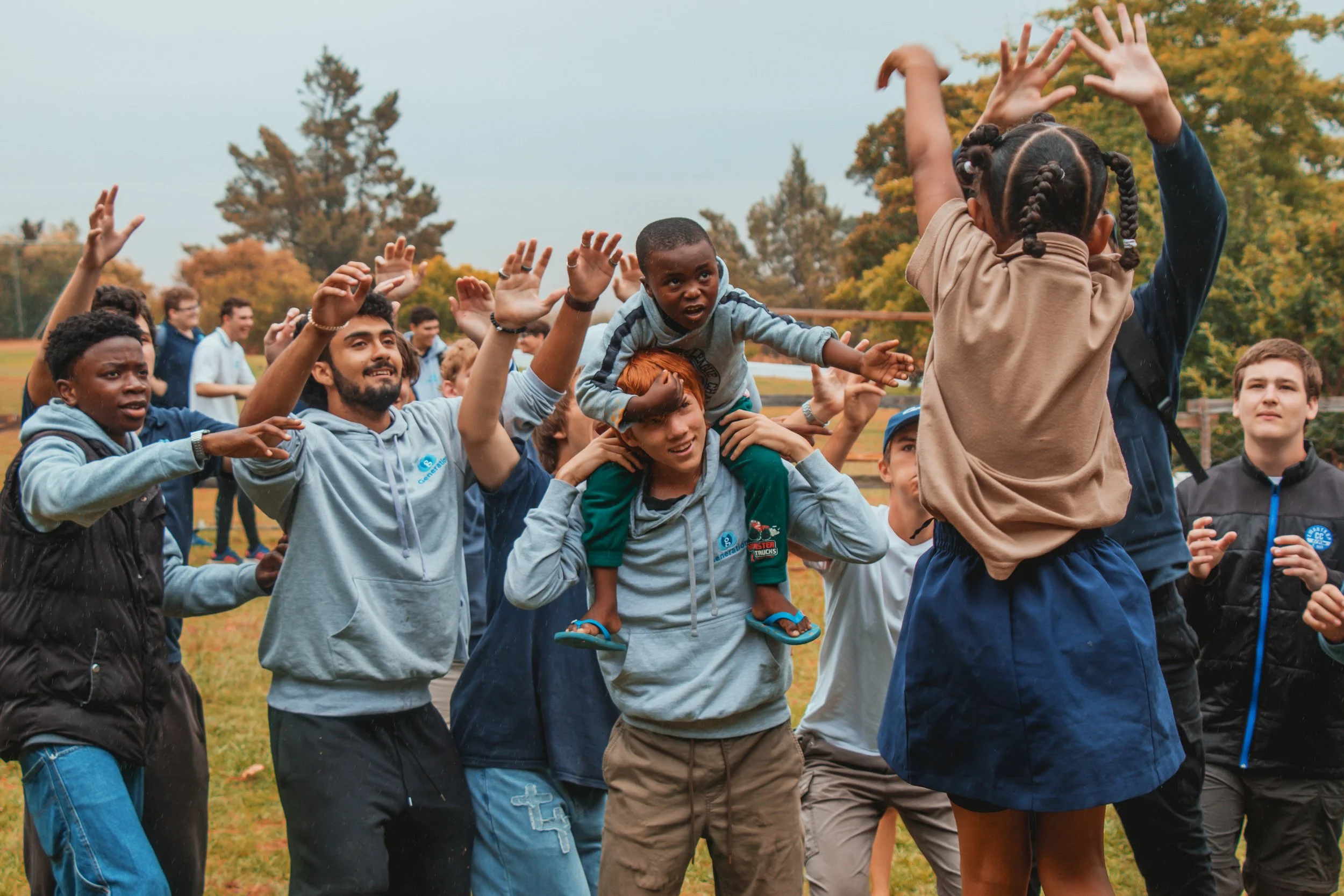 Group of children and young adults outdoors, participating in a team activity, with some standing and others sitting or being carried, in a park with trees and grass.