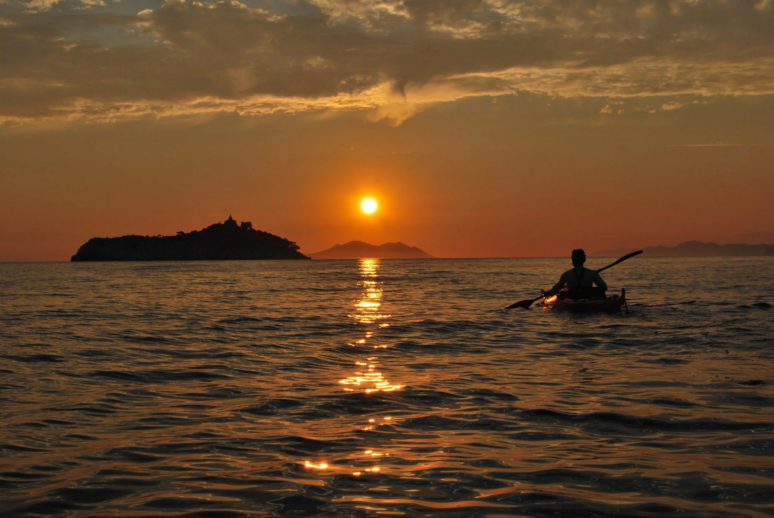 A person paddling a kayak on a calm body of water during sunset, with an island and mountains in the distance and a partly cloudy sky overhead.