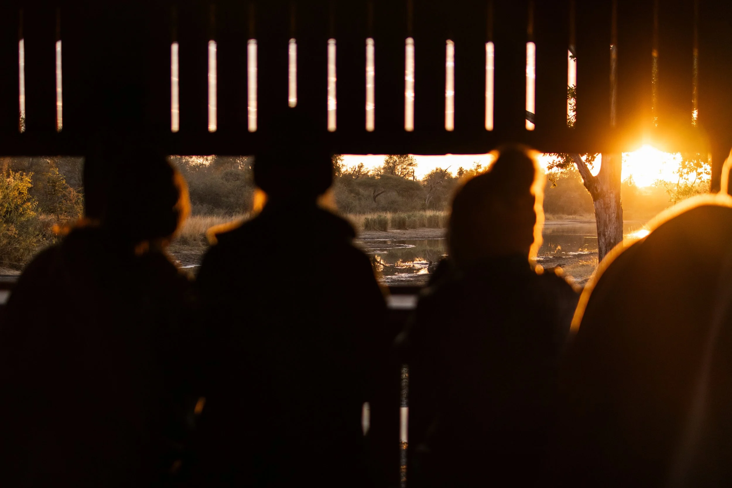 Silhouettes of people sitting under a wooden structure at sunset by a river.