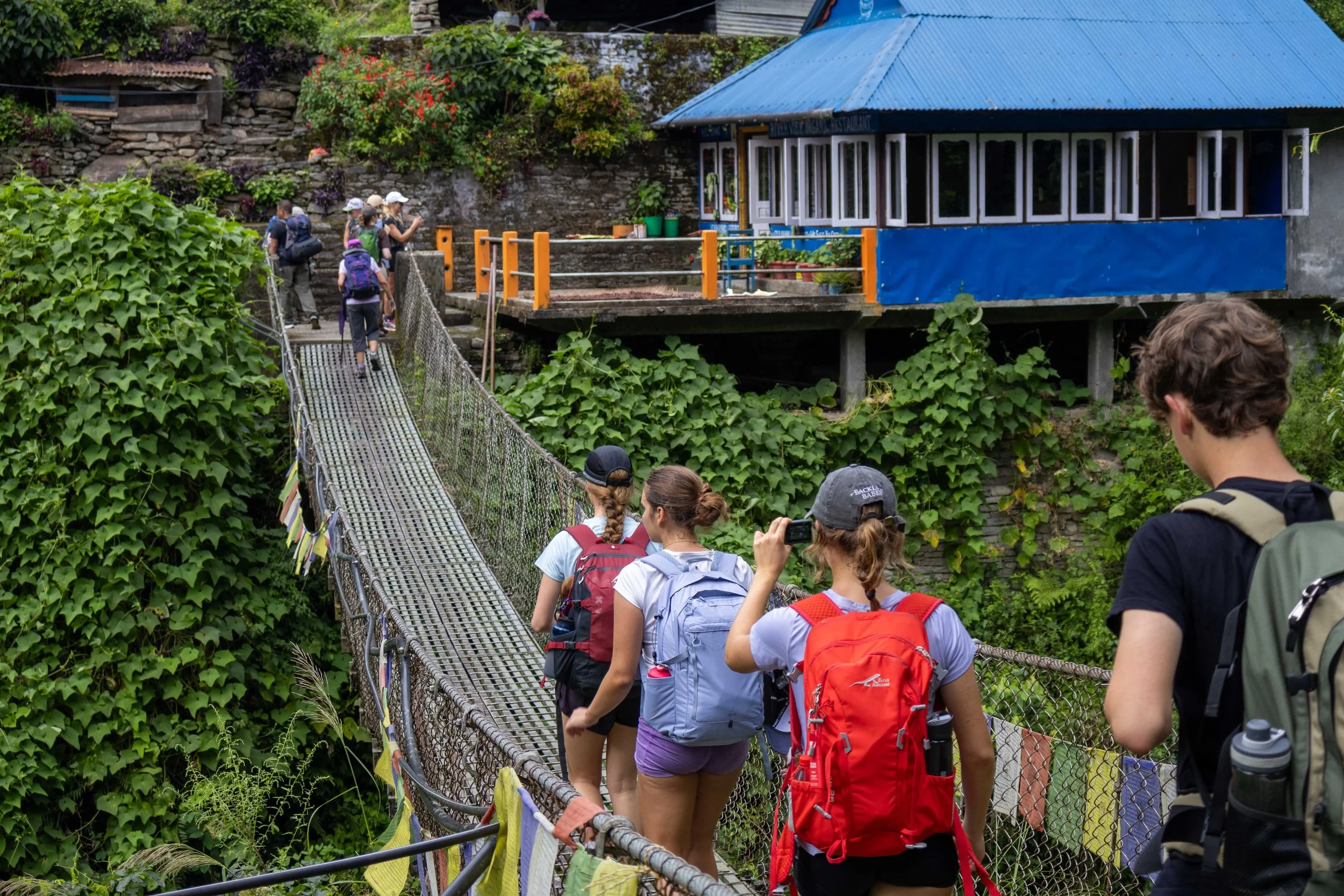 Group of hikers crossing a hanging bridge with a metal grate platform and railings, leading to a house with a blue corrugated metal roof, surrounded by lush green foliage and flowering plants.