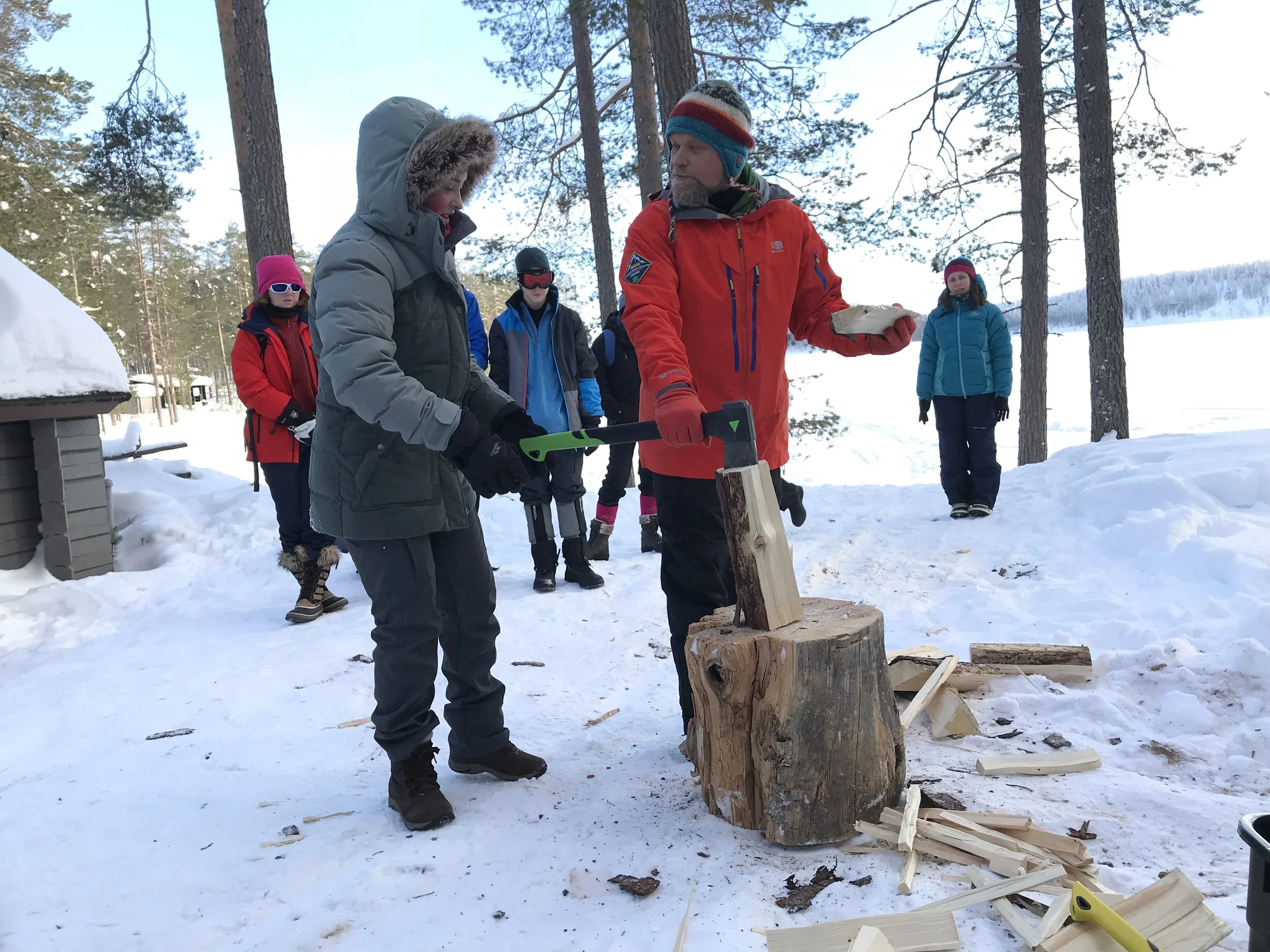 Group of people in winter clothing standing outdoors in a snowy forested area, around a tree stump being used as a chopping block for chopping wood.