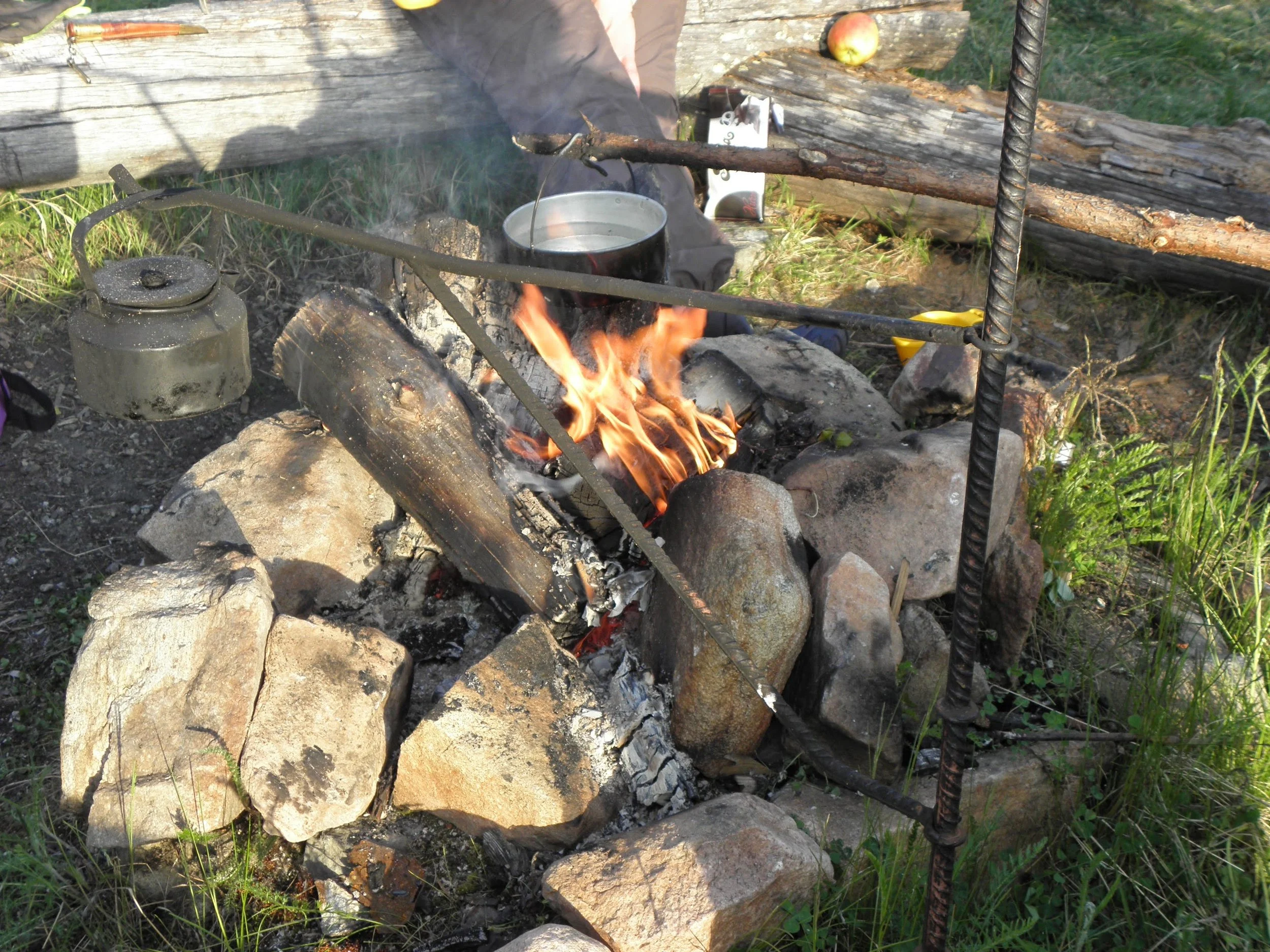 A fire burning in a campfire with rocks around it, set outdoors in a grassy area. There is a metal kettle on the left side of the fire, and a person with a mug sitting nearby. A fallen log and an apple are visible in the background.