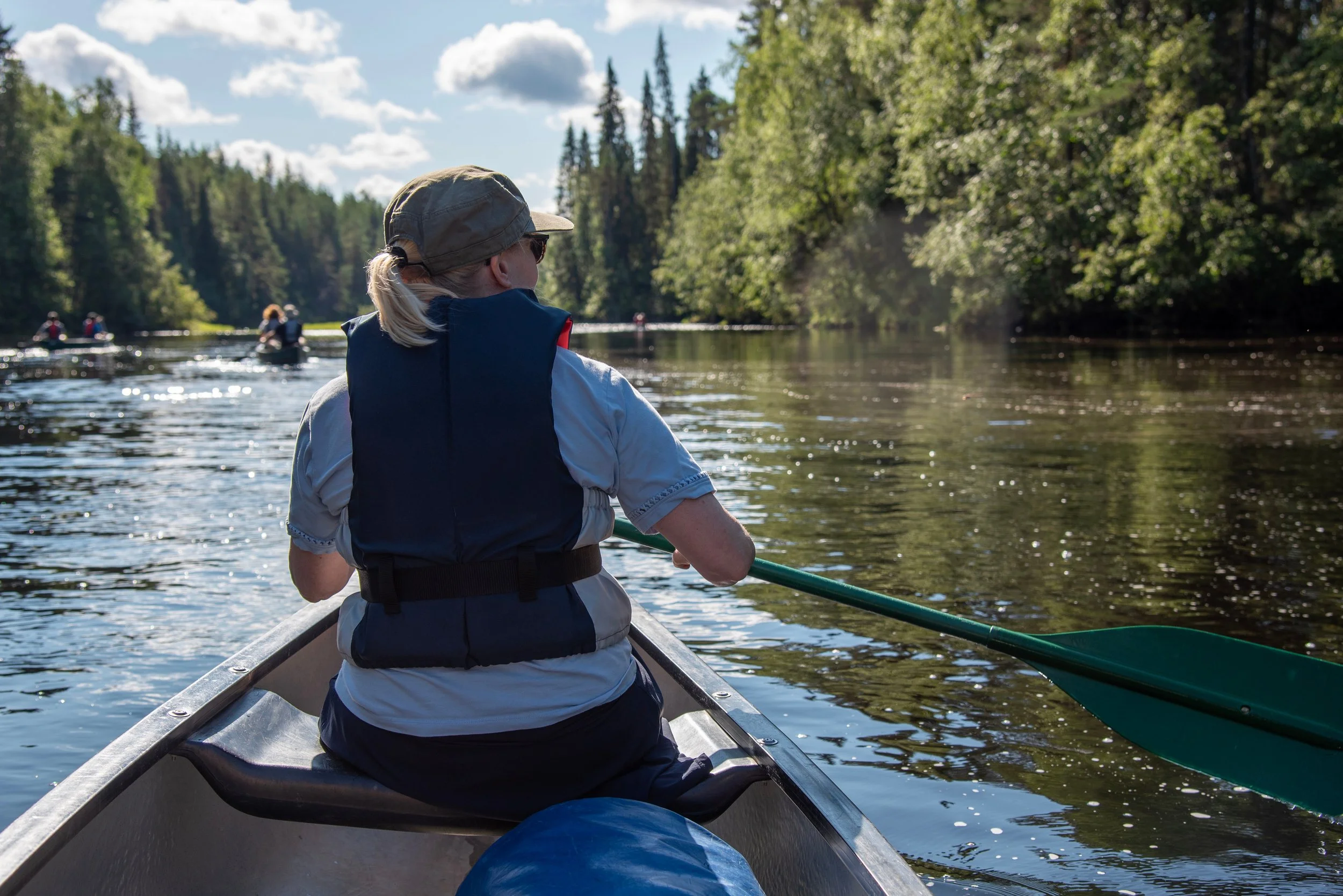 A person in a life jacket and cap paddling a canoe on a river surrounded by trees on a sunny day.