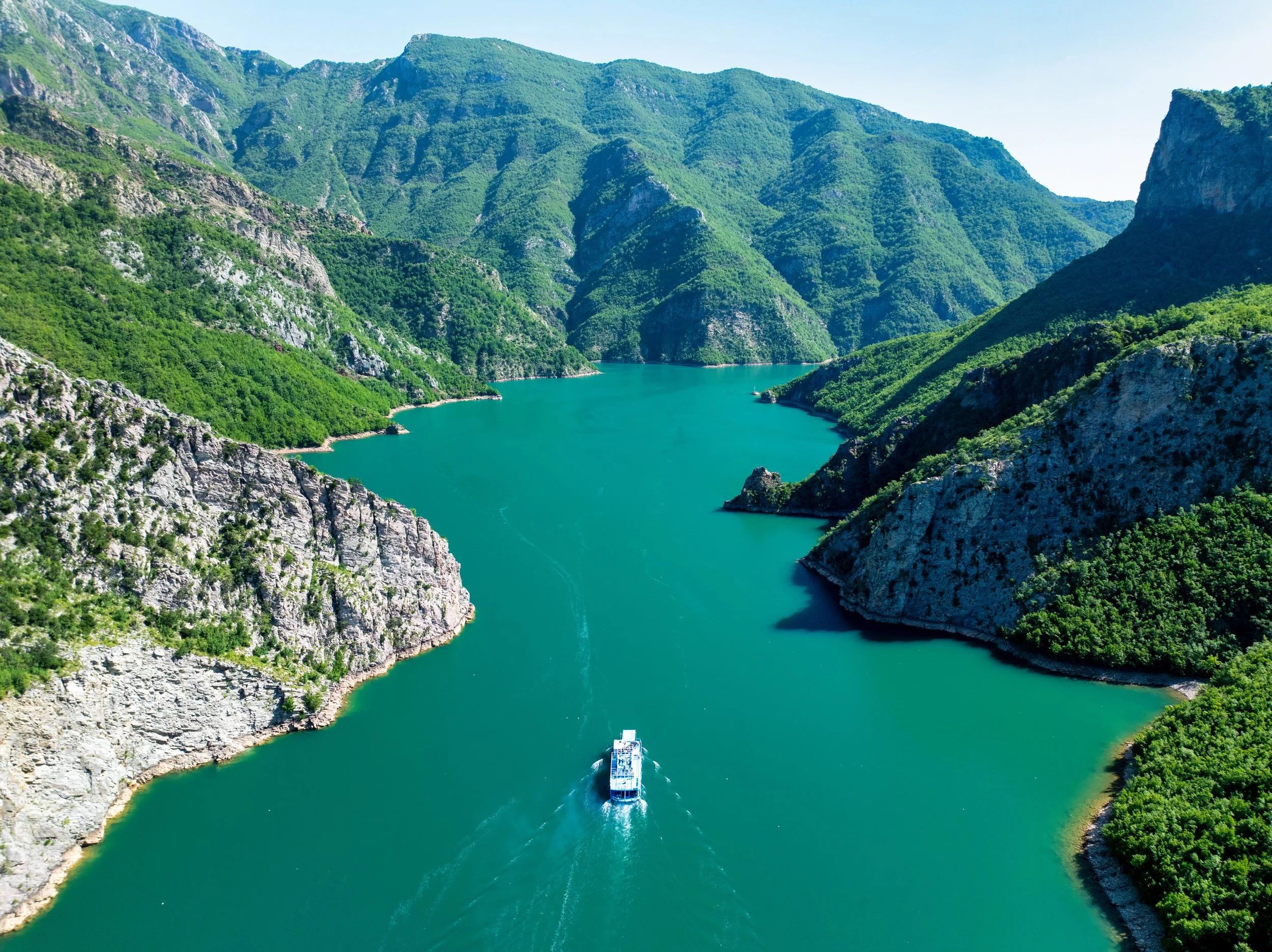 A boat sailing through a deep green river flanked by steep, lush green mountains on both sides under a clear blue sky.