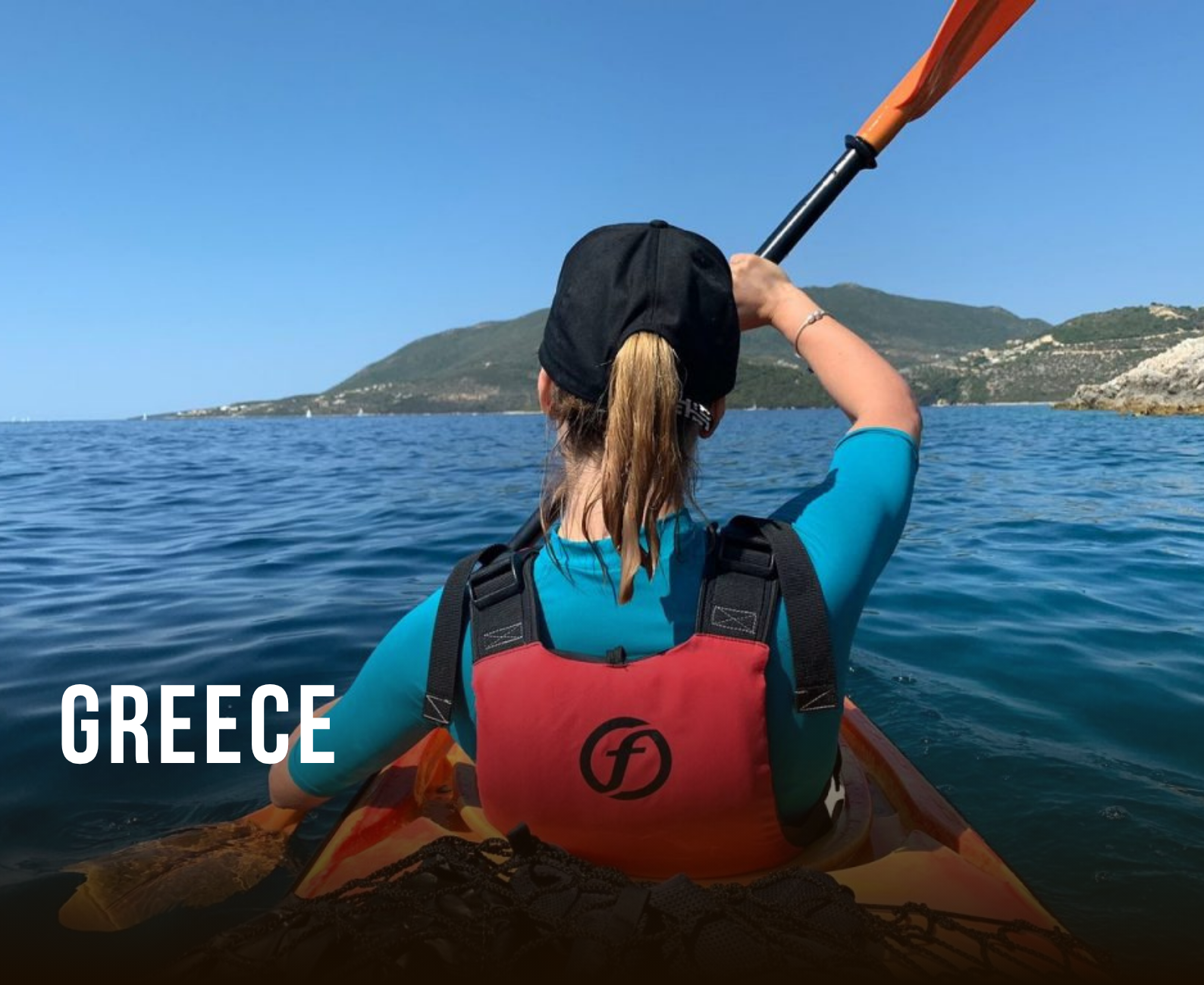 A person kayaking in Greece, wearing a black cap, a blue shirt, a red life jacket with a black logo, and holding a paddle, with a scenic view of mountains and water under a clear blue sky.