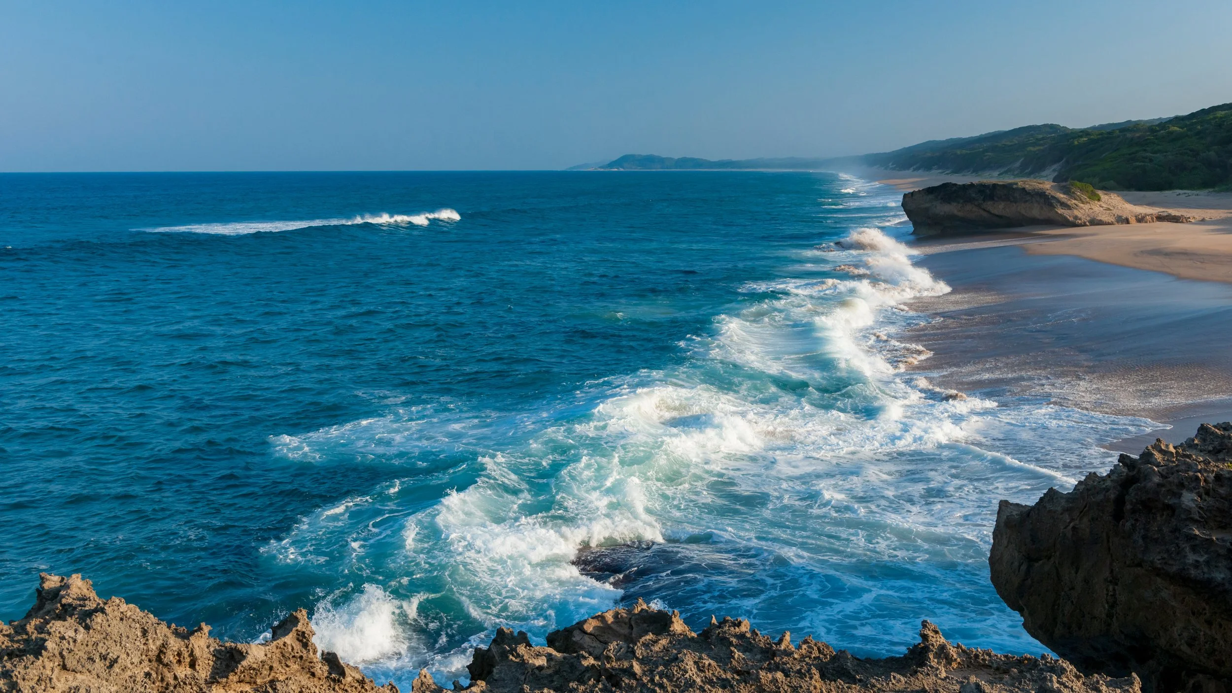 Ocean waves crashing against rocky shore with sandy cliffs in the distance under clear blue sky