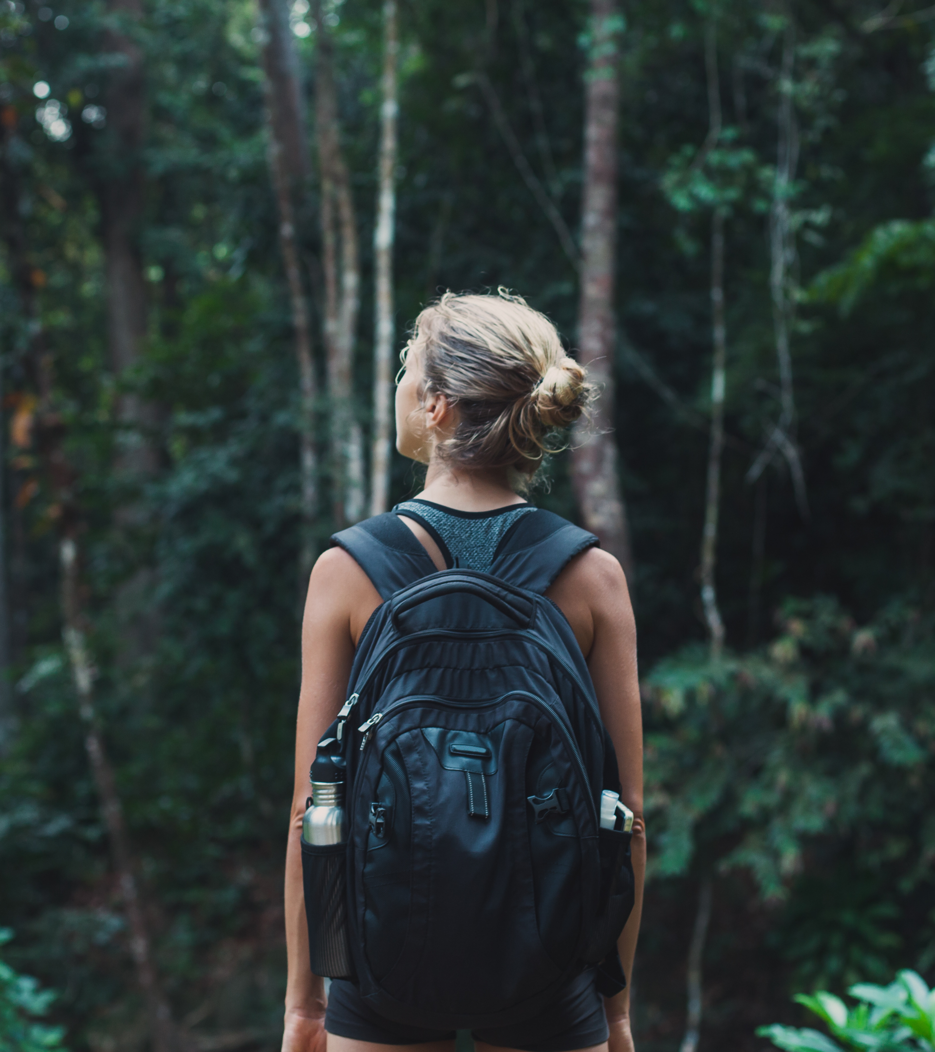 Woman with blonde hair tied in a bun, wearing a black tank top and a backpack, hiking through a dense forest.