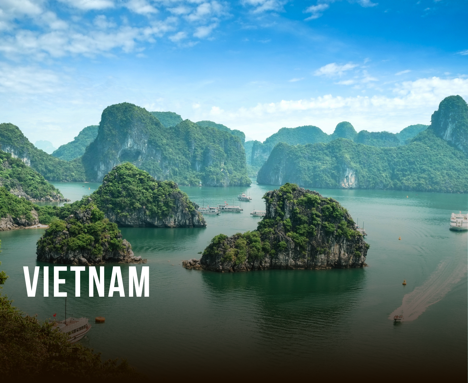 Scenic view of limestone islands and green hills in Halong Bay, Vietnam, with boats on calm waters and a bright blue sky with some clouds.