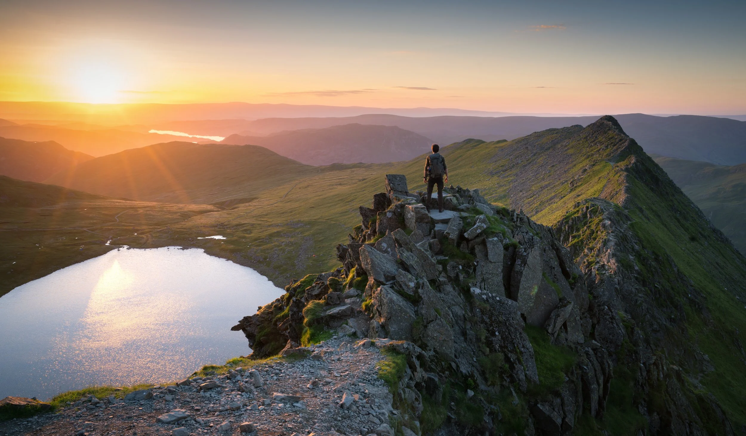 A hiker standing on a rocky mountain ridge at sunrise, overlooking rolling green hills and a lake below.