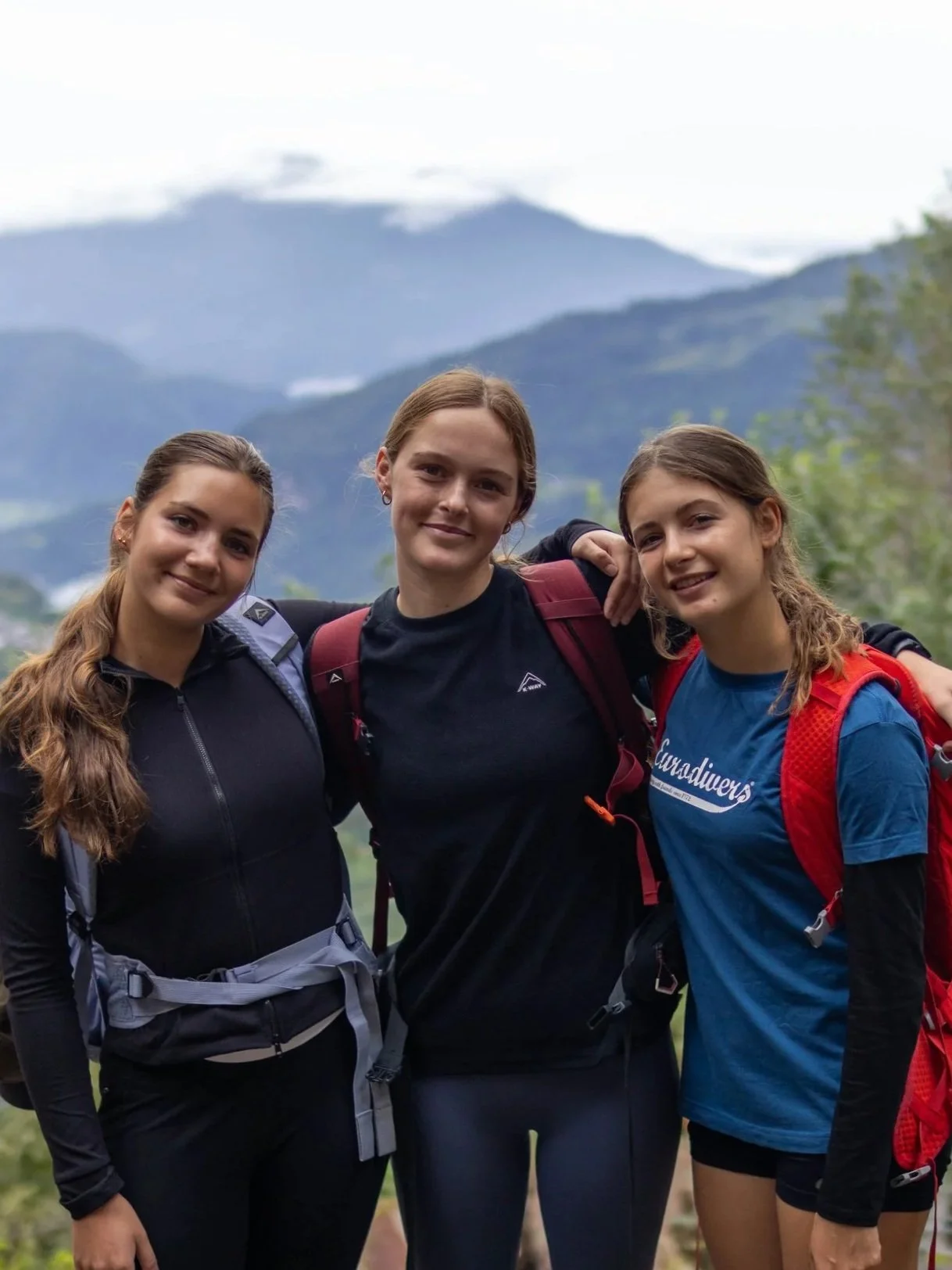 Three young women with backpacks smiling outdoors with mountains in the background.