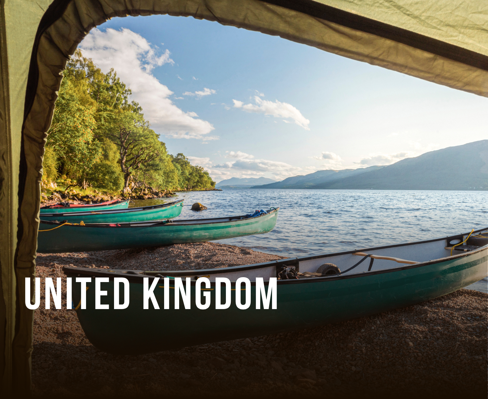 View from inside a tent looking out onto a lake with kayaks on the shore, green trees along the shoreline, and mountains in the distance under a partly cloudy sky.