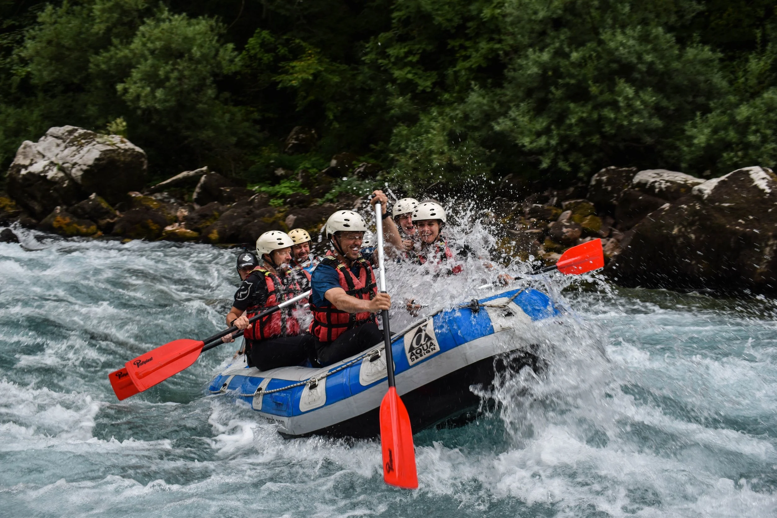 Group of people white water rafting on a river, wearing helmets and life jackets, with some holding paddles, surrounded by rocks and dense green trees.