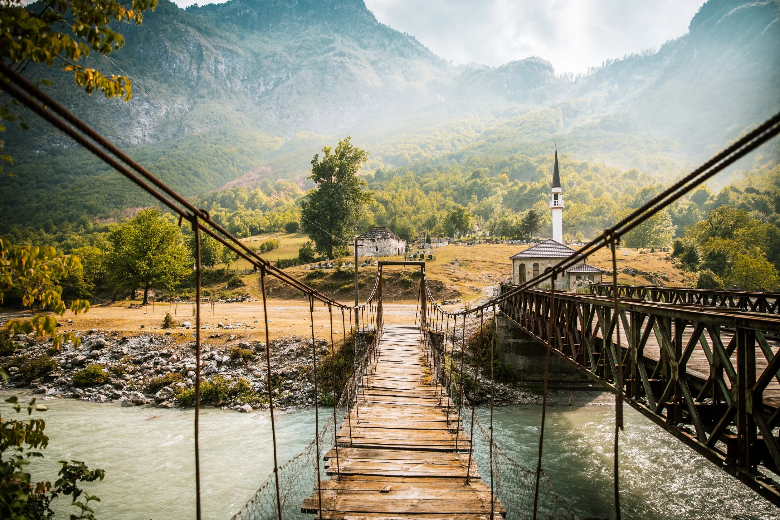 A wooden suspension bridge crossing a river with mountains and a church with a tall steeple in the background.