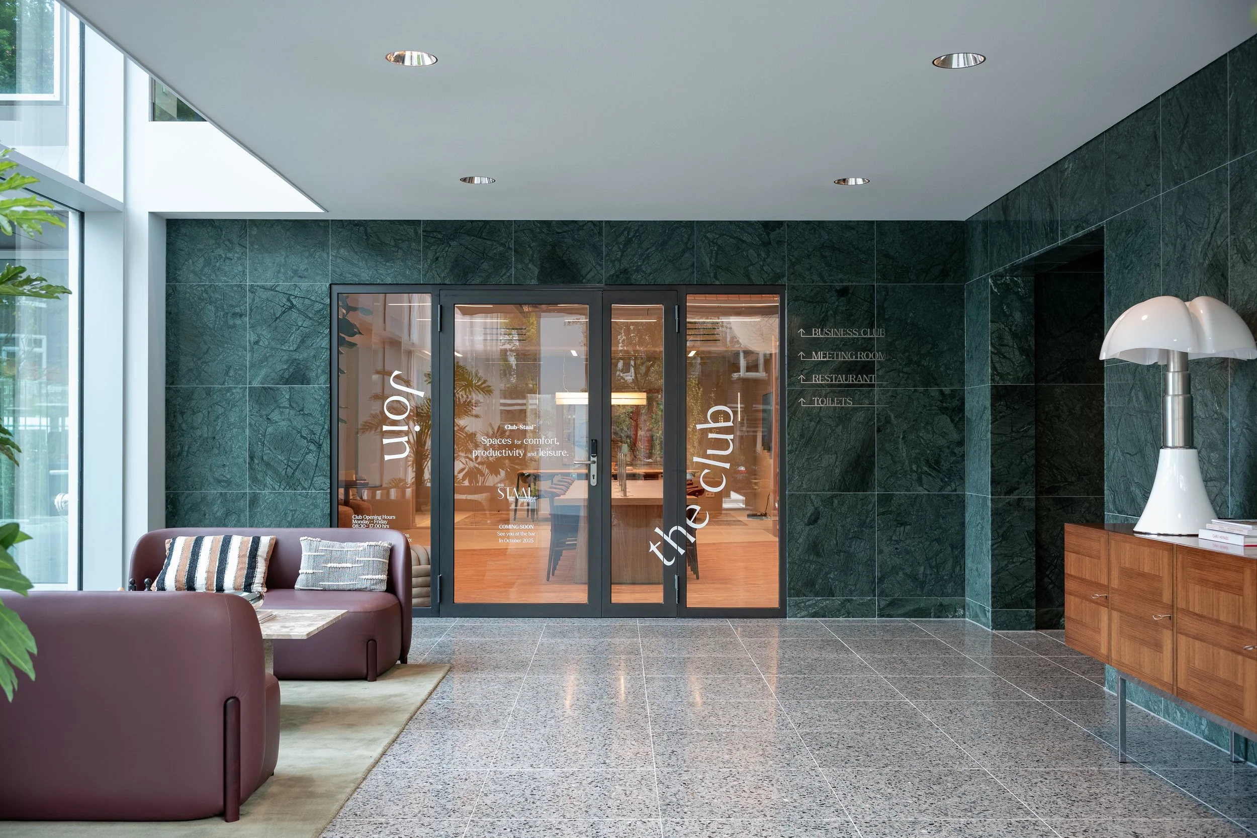 Lostaankamer in een moderne lobby met groene marmeren muur, een bankenbank met kussens, een houten console met een groot witte lamp en een deur met de tekst 'theclub' en raampanelen die het interieur van de club zichtbaar maken.
