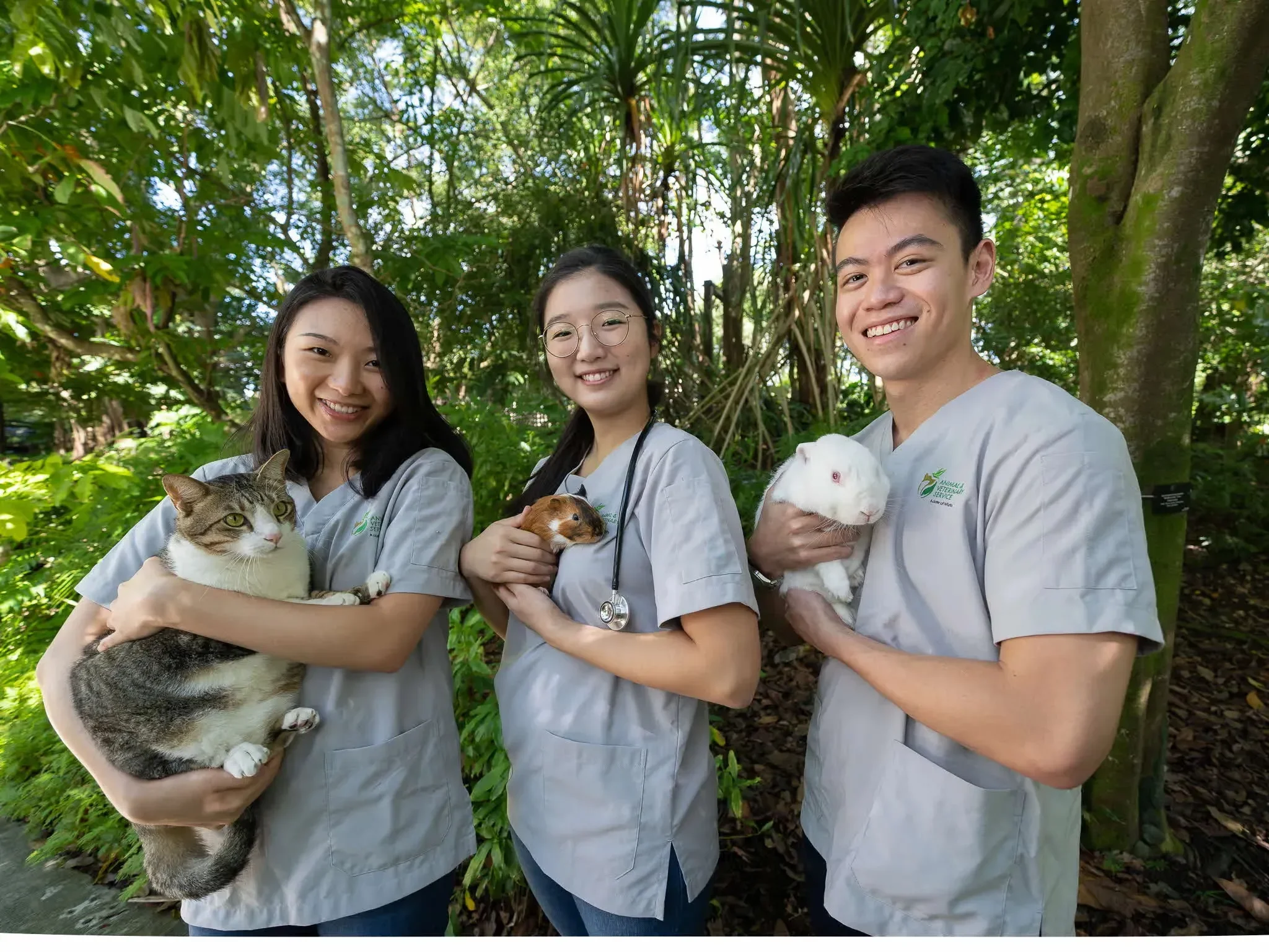Three young Animal Medical Center veterinarians smiling outdoors in a lush, green forest, each holding a different small animal: a cat, a guinea pig, and a rabbit.