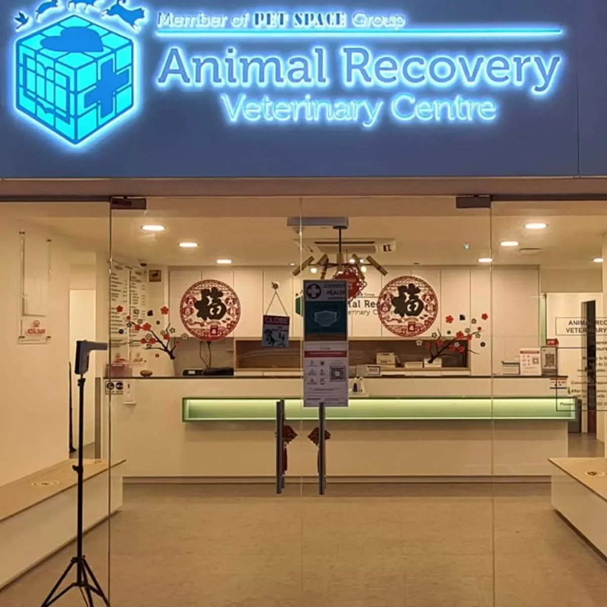 The entrance of Animal Recovery Veterinary Centre with a neon sign displaying its name and a logo of a blue cube with a cross and a heart. Inside, there are decorative red and black Chinese-style paper cutouts on the glass, a reception counter with a green light underneath, and some informational posters and signs including a stand with a face mask notice.
