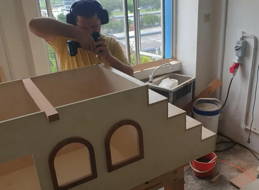 A person wearing yellow, working on a woodworking project with a drill, standing behind a partially constructed wooden structure with arched cutouts, inside a workshop or garage with a window revealing an outside view of parking lot and trees.