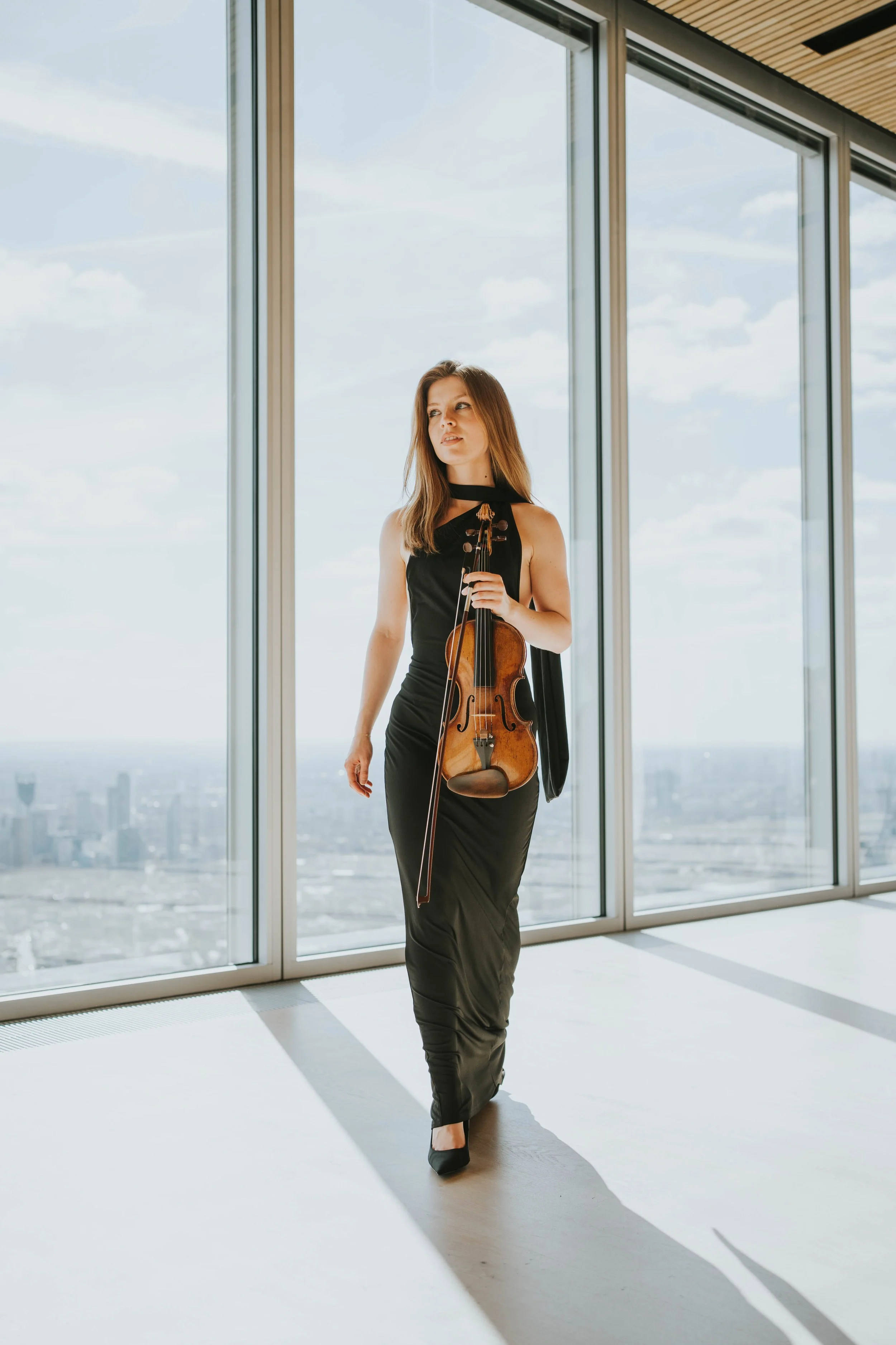 A woman in a black evening gown holding a violin, standing in front of large windows in a high-rise building, with city skyline visible outside.