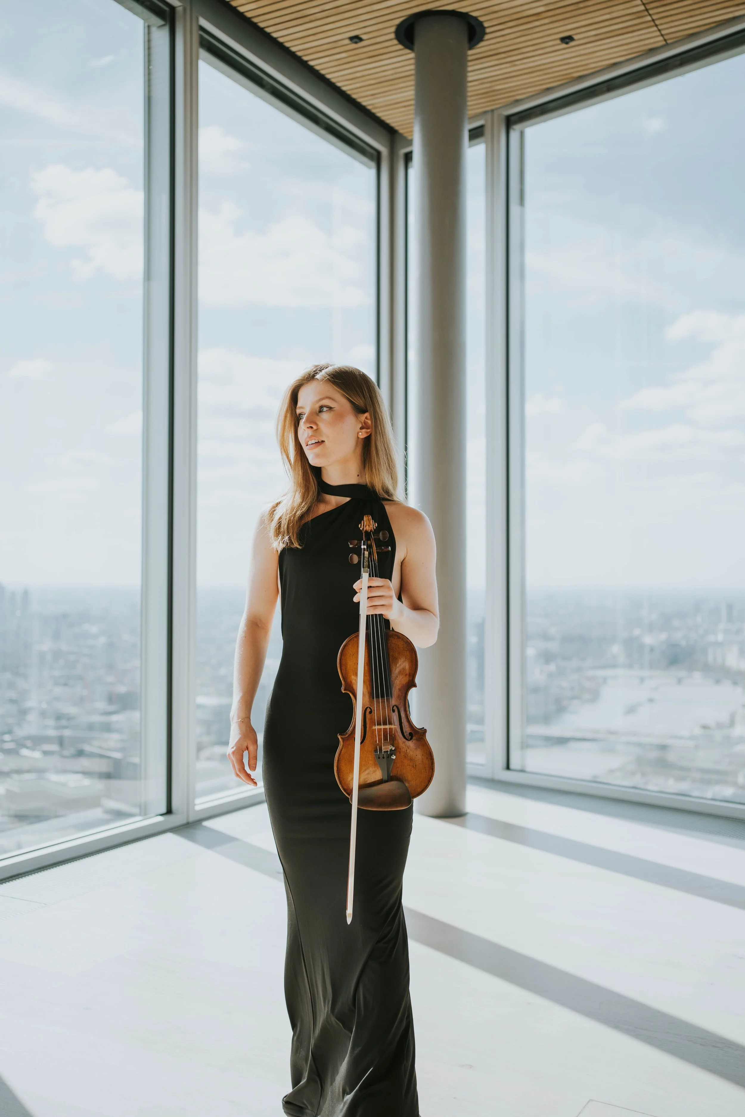 A young woman in a black gown holding a violin stands in a modern room with large floor-to-ceiling windows overlooking a cityscape under a cloudy sky.