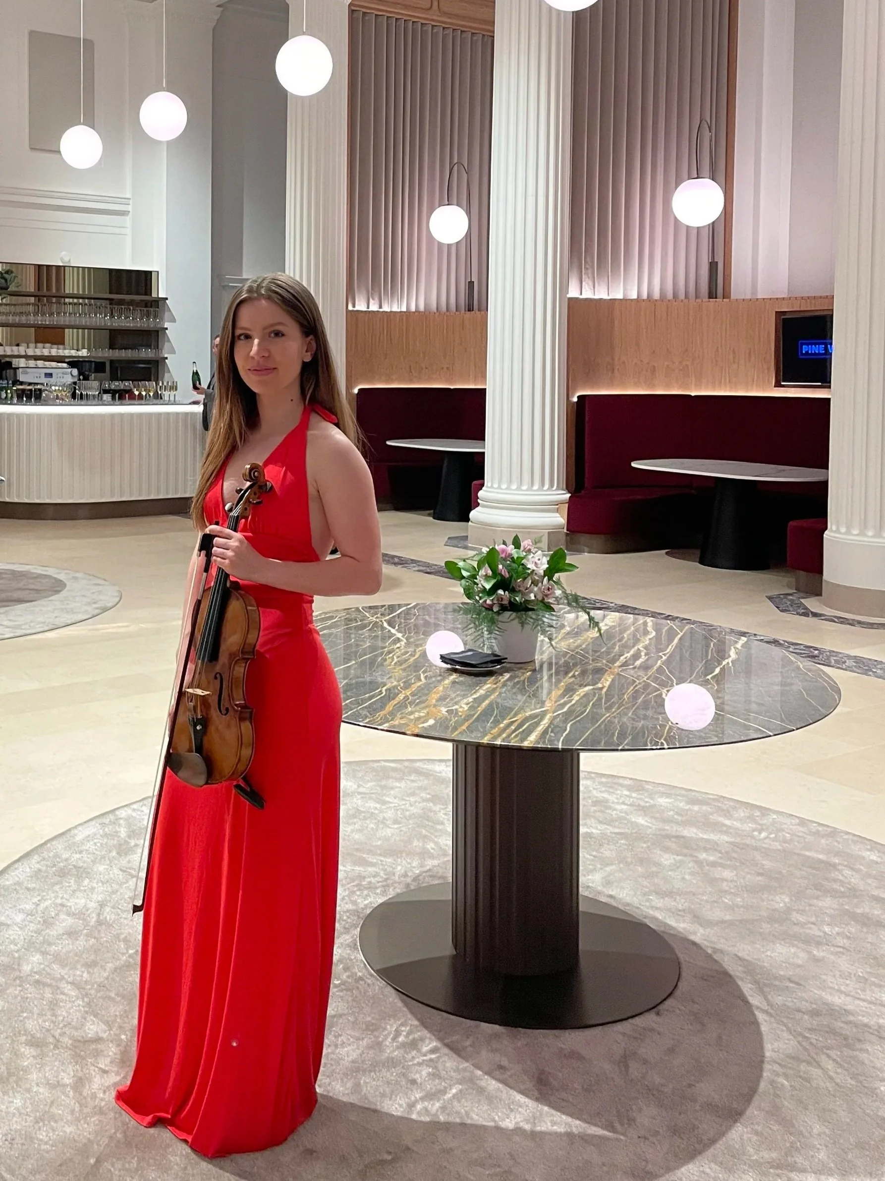 A woman in a long red dress holding a violin, standing in a stylish, modern interior space with white pillars, round hanging lights, a marble-top table with a flower arrangement, and a seating area with red booths in the background.