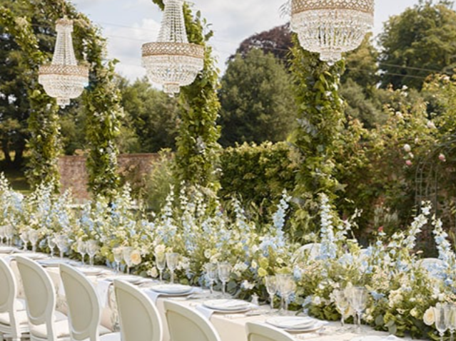A long outdoor dining table decorated with white and light blue floral arrangements, surrounded by white chairs, with crystal chandeliers hanging above and green trees in the background.