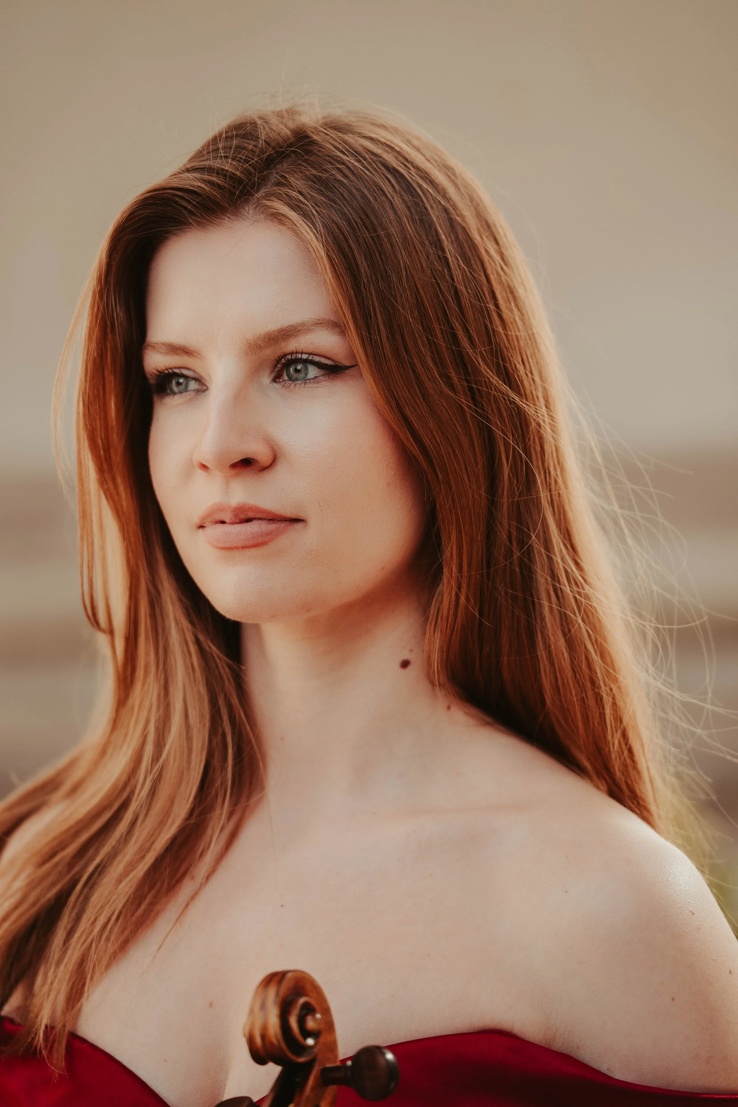 A young woman with long red hair and blue eyes, looking thoughtfully to the side, with a blurred background and a visible cello's scroll in the foreground.